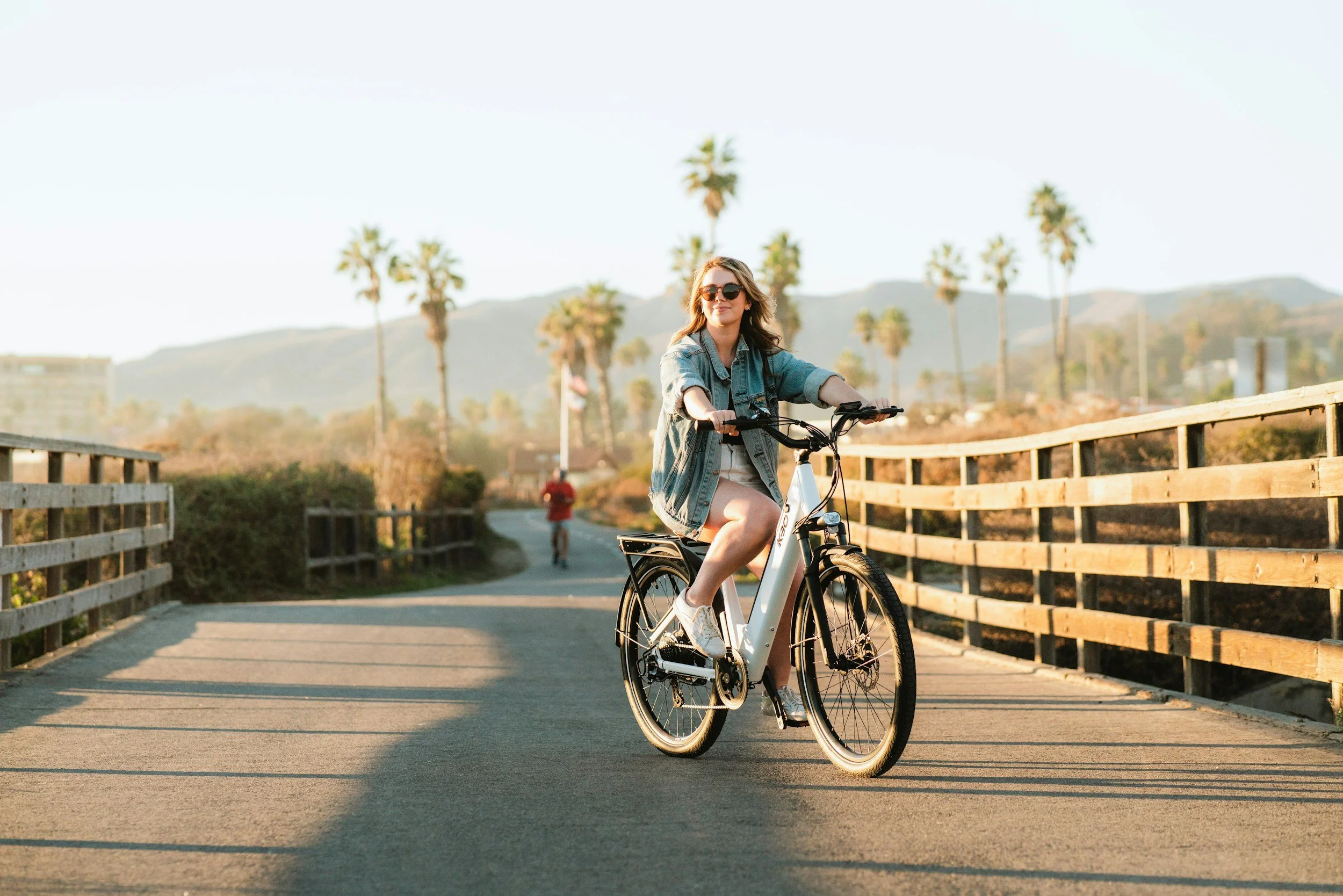 A woman wearing sunglasses and a denim jacket riding a bicycle on a bridge during sunset, with palm trees and hills in the background.