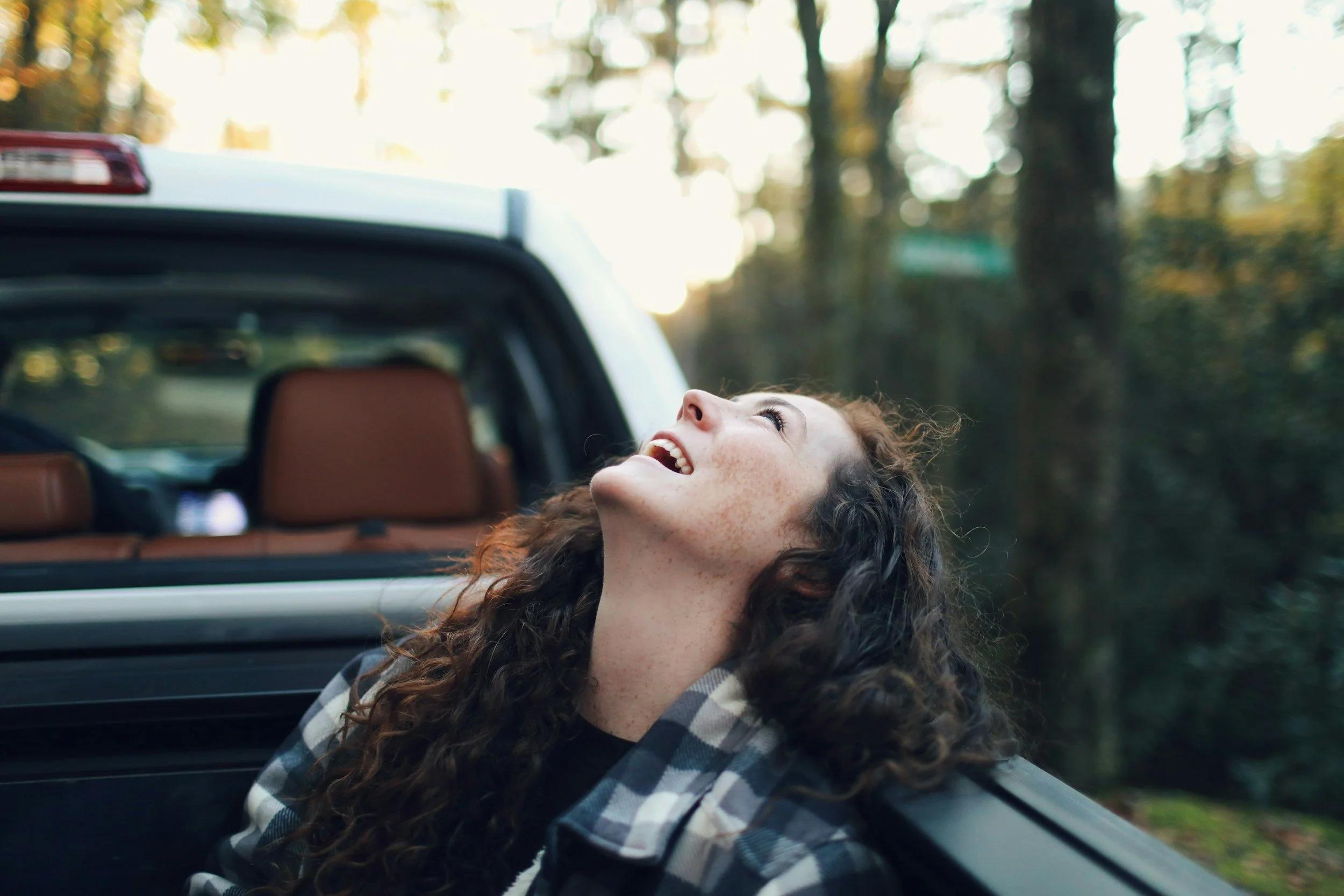 A woman with curly brown hair smiling and looking up while sitting in the back of a vehicle with the trunk open during autumn, with trees and sunlight in the background.