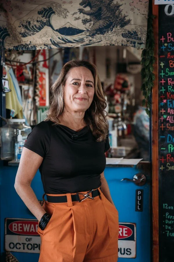A woman standing inside a colorful shop with a blue counter, chocolate and colorful chalkboard menu, and artwork hanging above her. She is wearing a black t-shirt and orange pants.