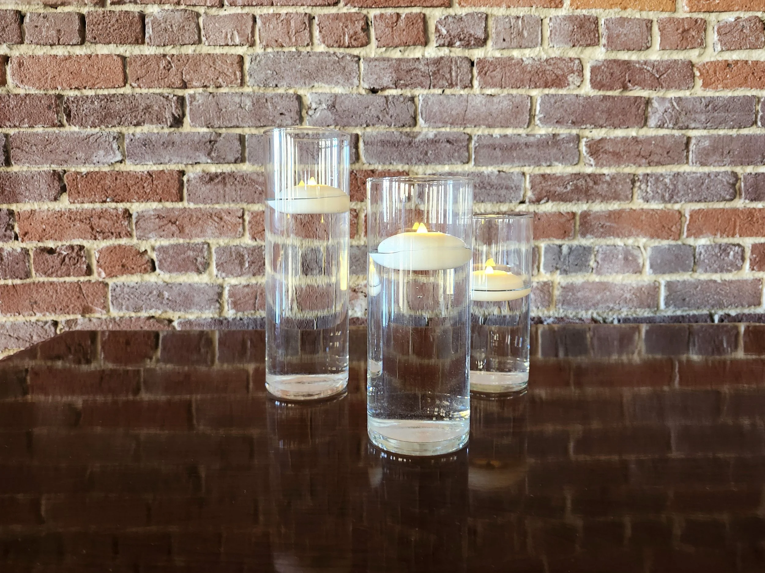 Three tall glass candle holders with floating candles on a wooden surface against a brick wall.