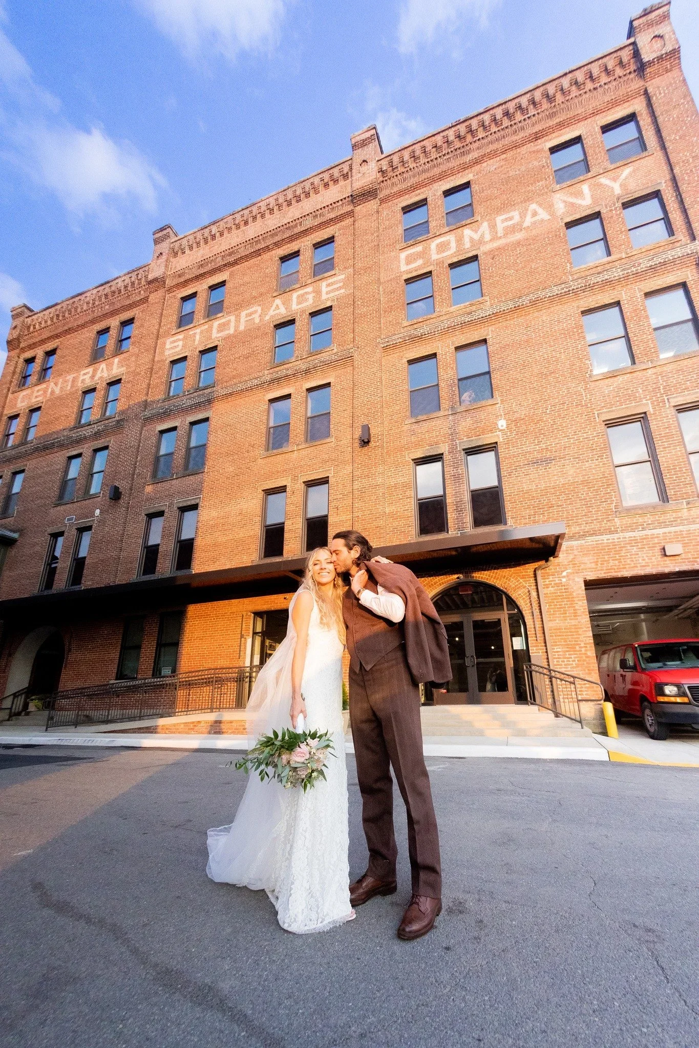 A newlywed couple standing outside a brick building with the words `Central Storage Company` written on it. The bride is in a white wedding dress holding a bouquet, and the groom is in a brown suit, kissing her on the cheek under a blue sky.
