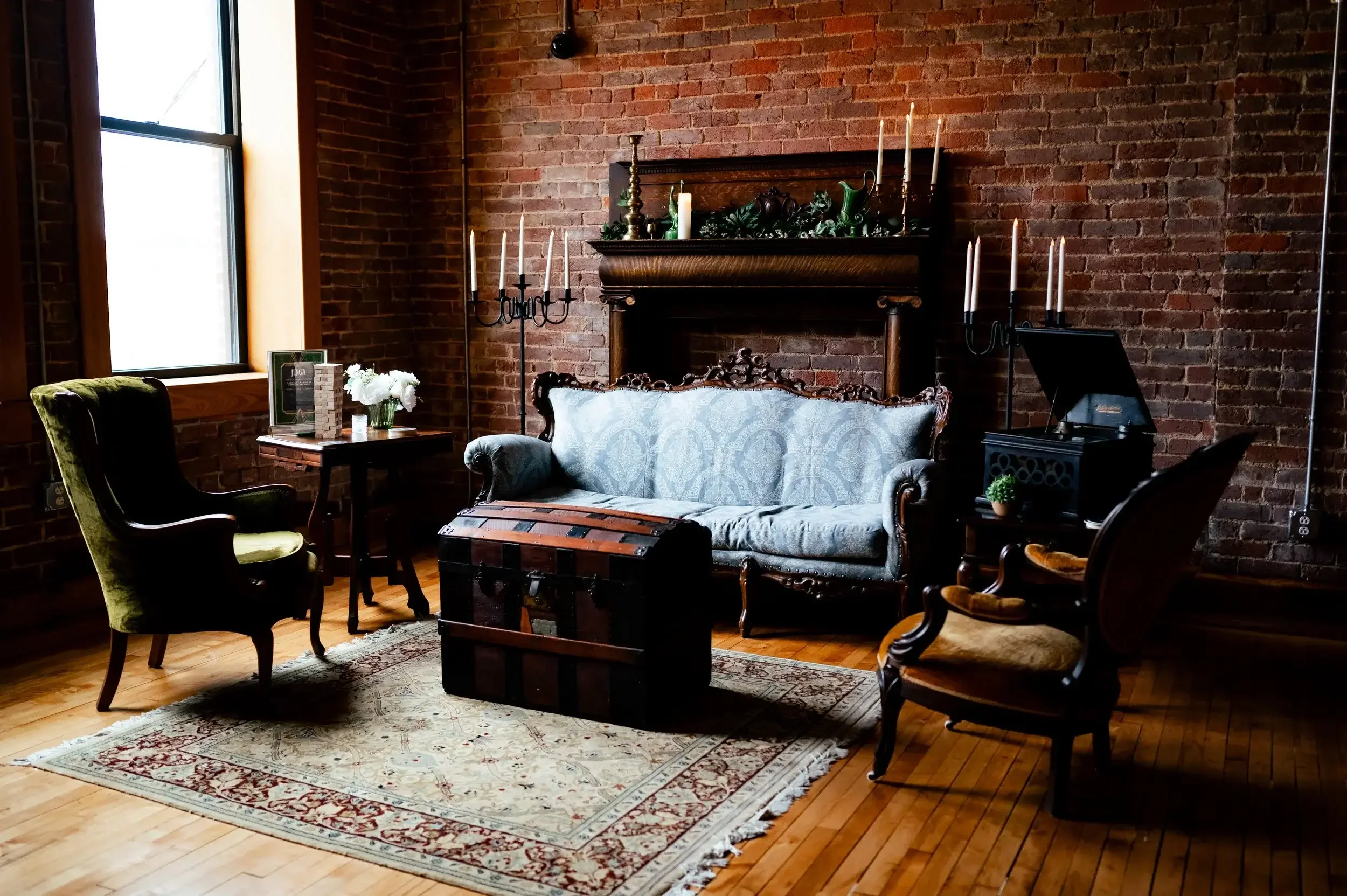 Interior of a vintage living room with brick walls, wooden floor, ornate sofa, two armchairs, a trunk, a small side table, a fireplace mantle with candles and greenery, and a painting on a side table, lit by natural light from a window.