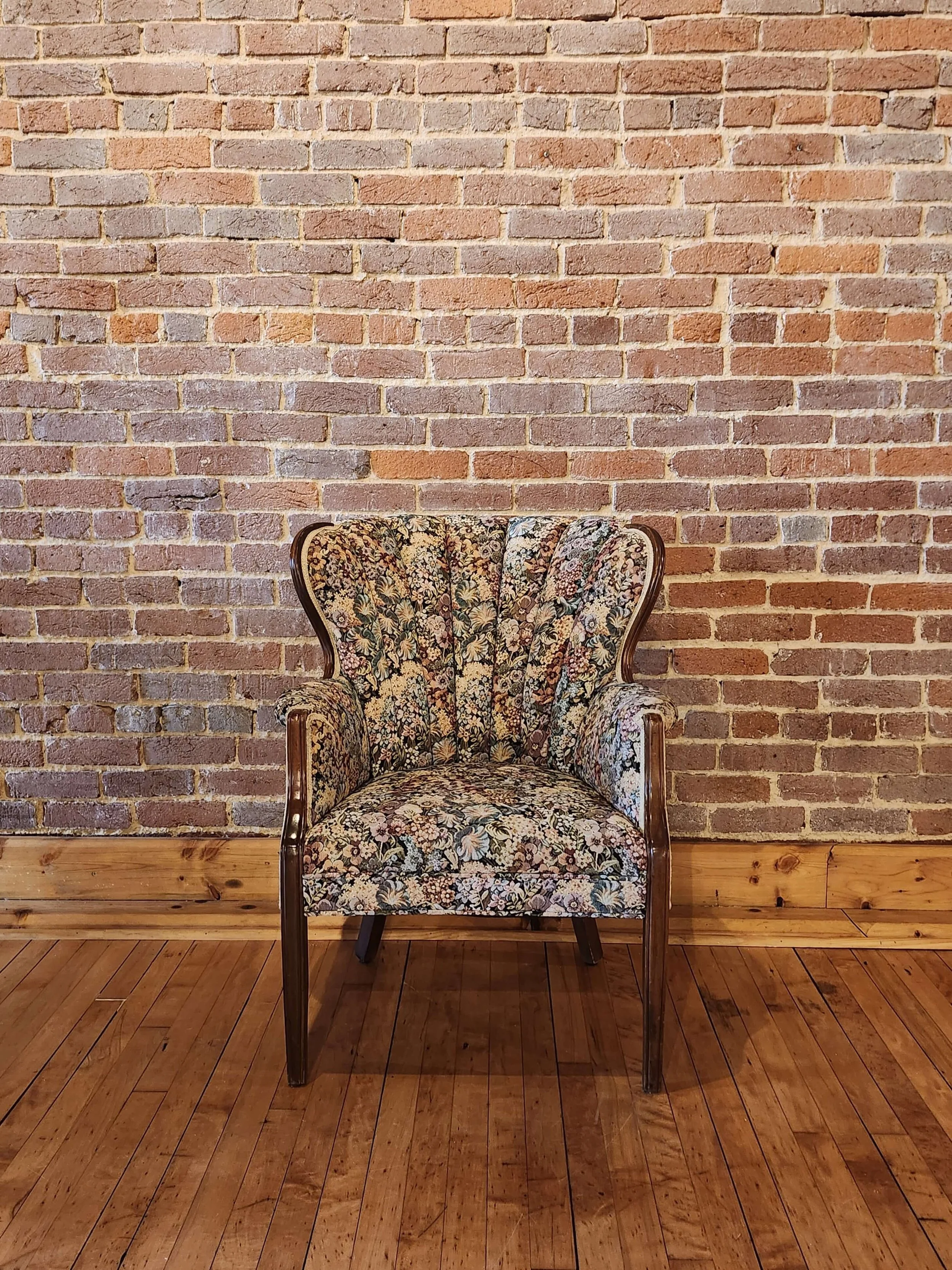 A floral upholstered armchair with wooden trim placed against a brick wall on a wooden floor.