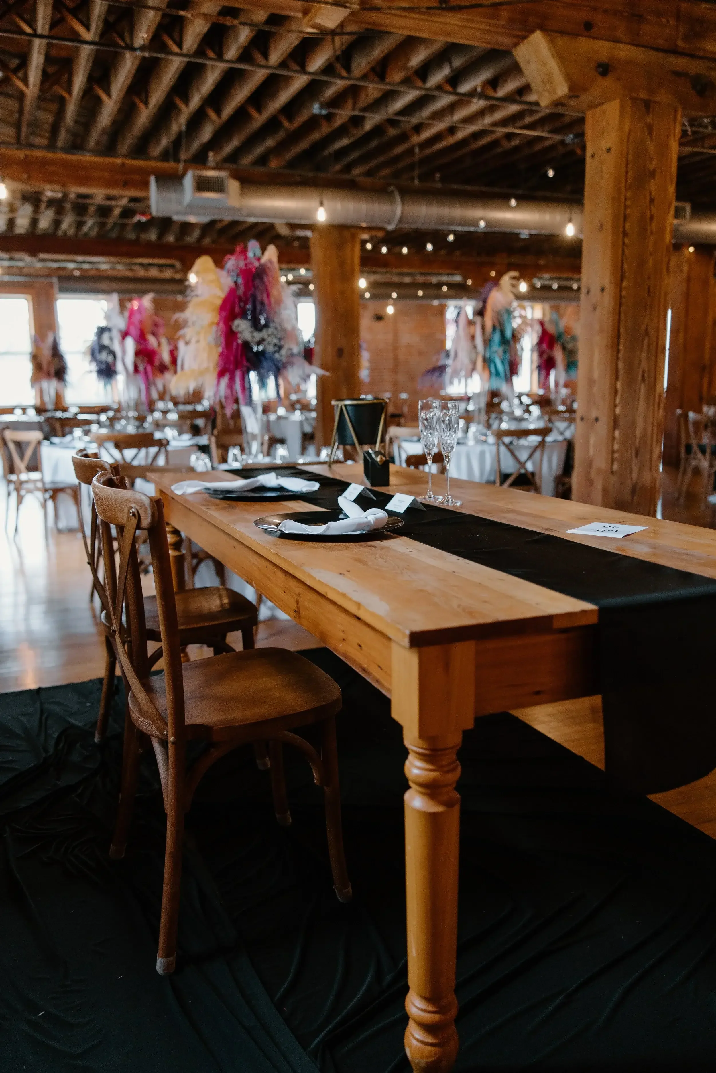 Wedding reception setup with a rustic wooden table, elegant tableware, and colorful feathered centerpieces, inside a warmly lit venue with exposed wooden beams.