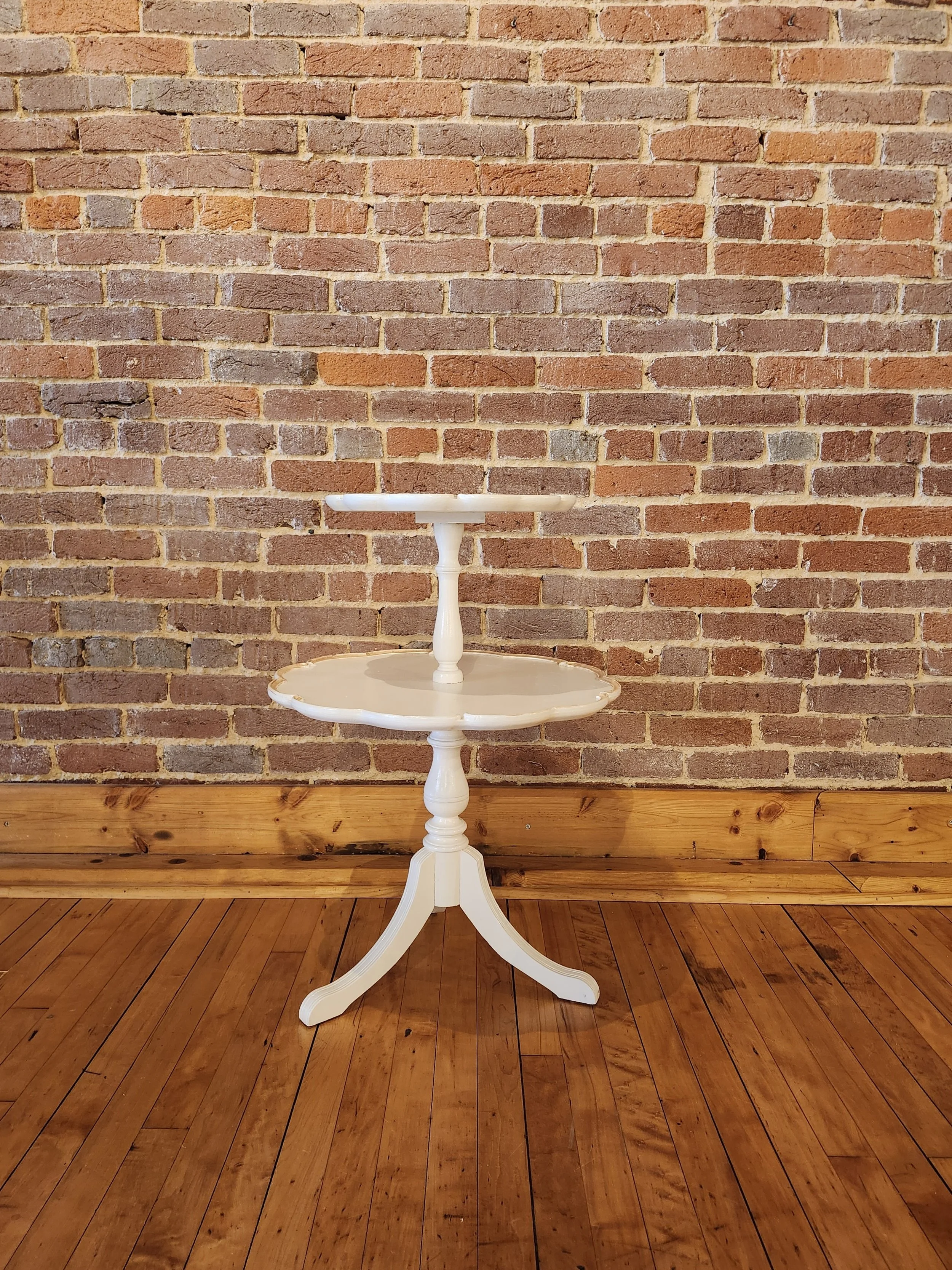 A white two-tiered round side table with turned legs, positioned against a brick wall and on a hardwood floor.