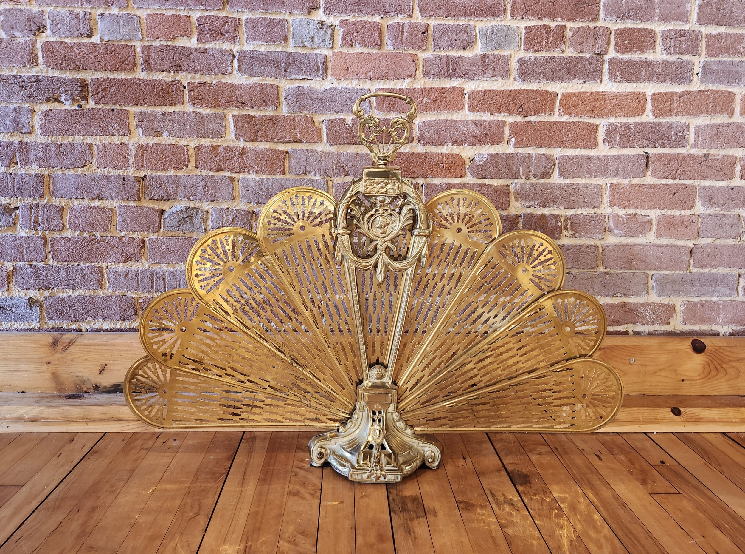 Ornate gold fireplace screen with intricate designs, leaning against a brick wall on a wooden floor.