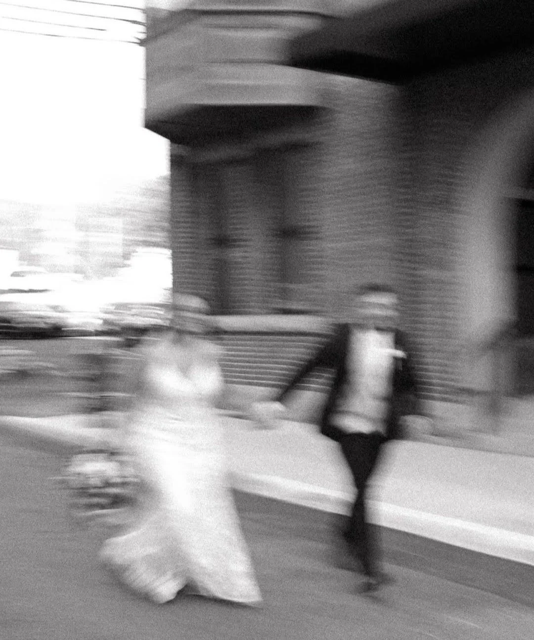 Black and white photo of two people walking on a city sidewalk near a brick building, one person carrying a plastic bag, motion blur visible.