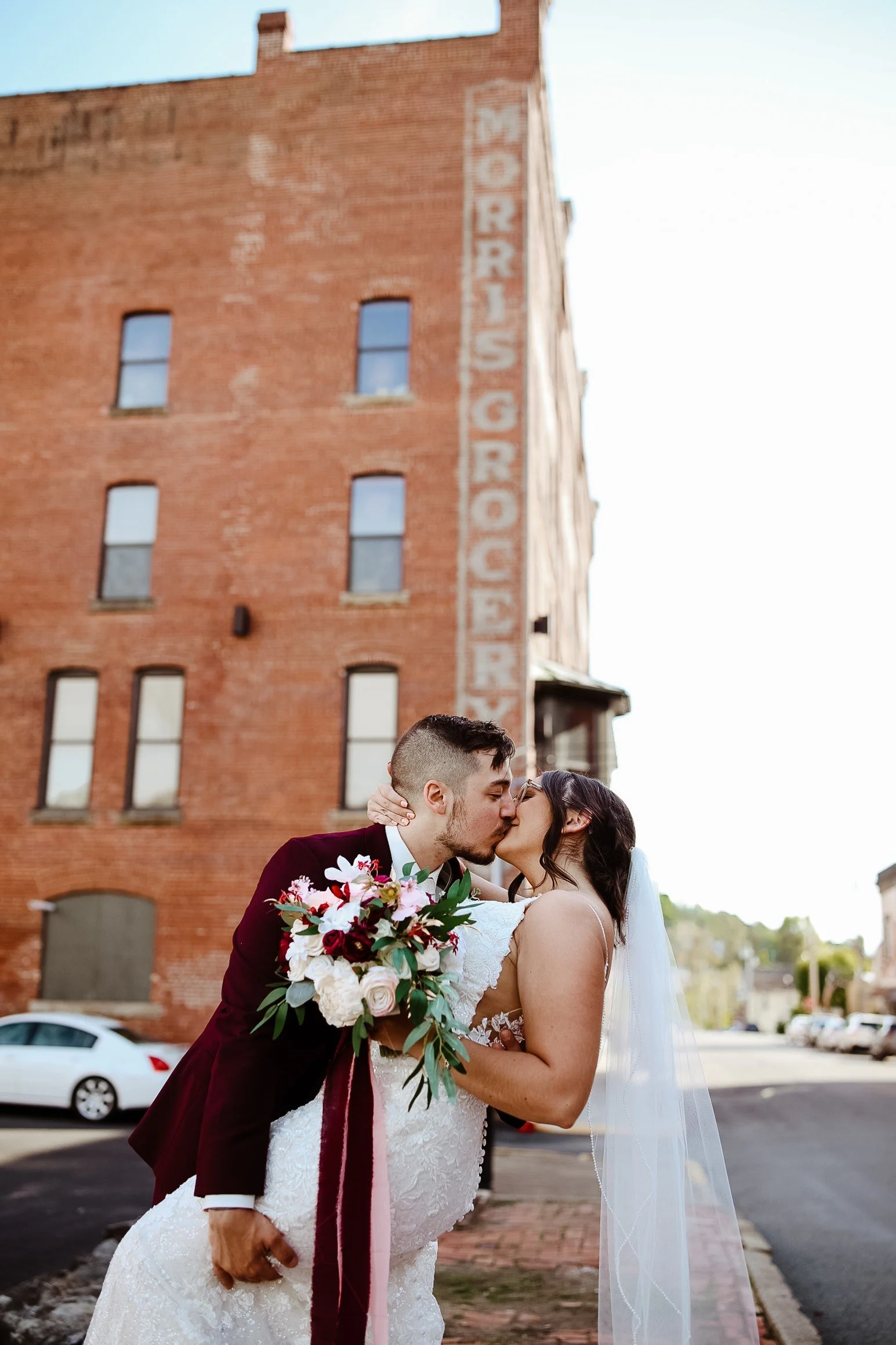 A newlywed couple shares a kiss on a city street, with the groom holding a bouquet of flowers and the bride wearing a white wedding gown and veil. An old brick building with a faded sign reading 'Morris Grocery' is visible in the background.