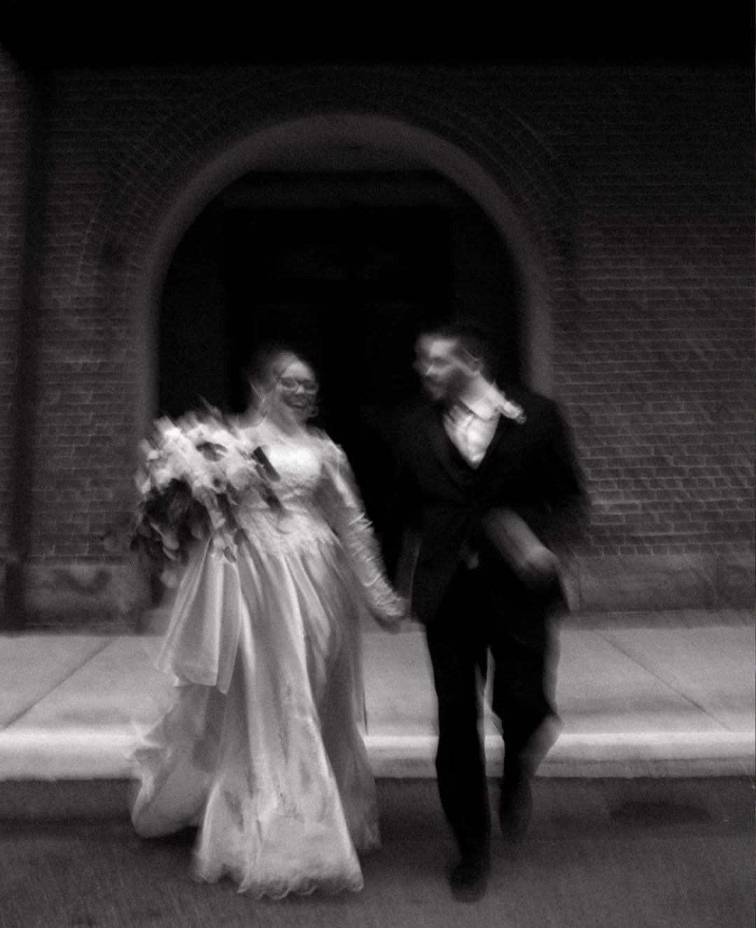A couple in wedding attire walking hand in hand outside a brick archway, smiling and enjoying the moment.