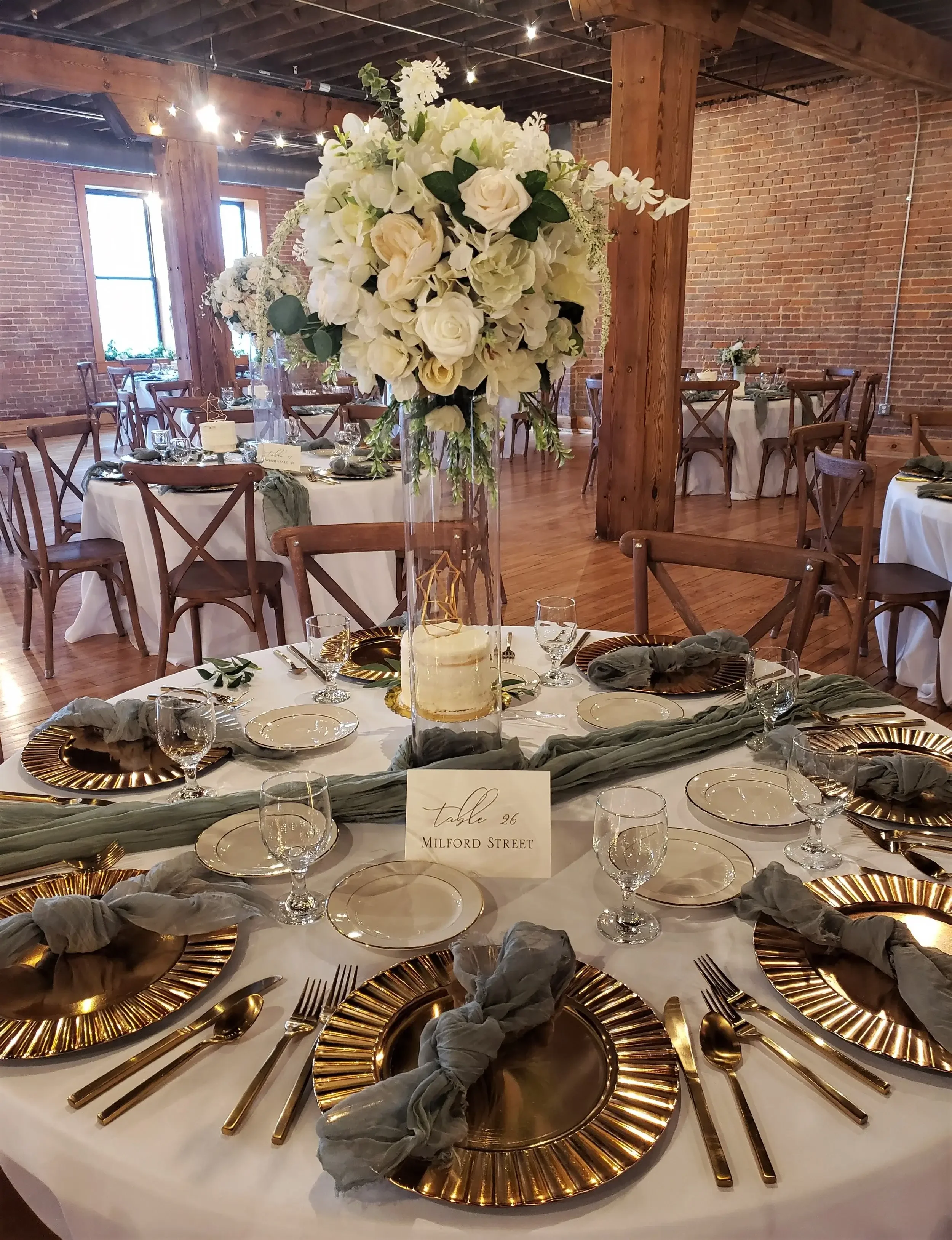 Elegant wedding reception table with a tall floral centerpiece of white roses and greenery, gold-rimmed chargers, gray napkins tied with string, clear glassware, and a white tablecloth in a rustic venue with brick walls and wooden beams.