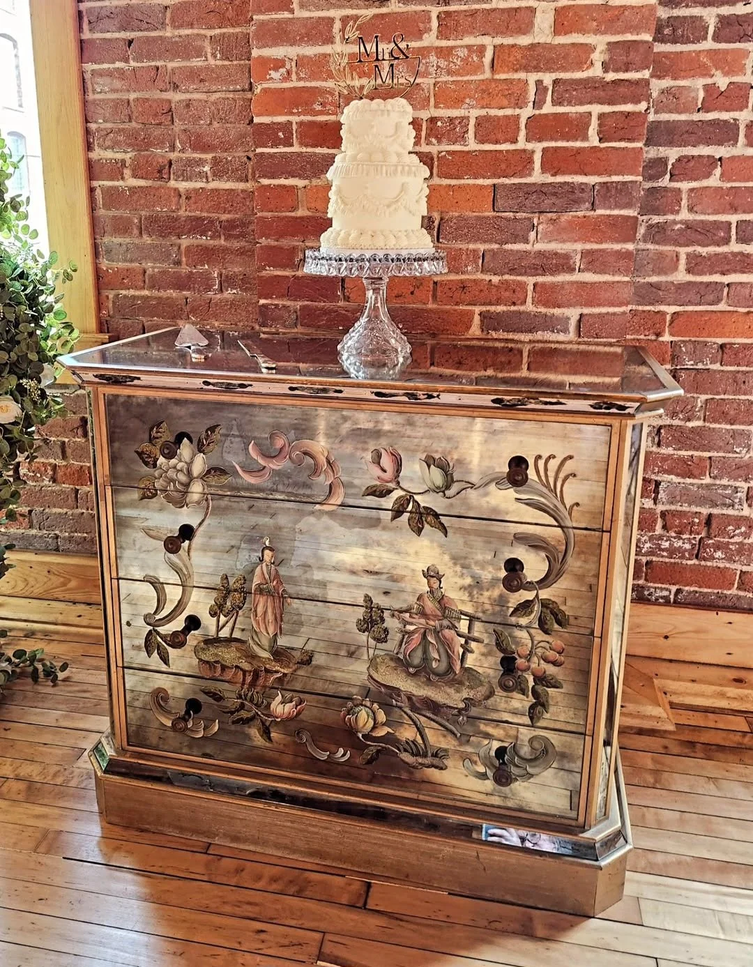 A vintage wooden cabinet with painted floral and figurative designs, topped with a glass cake stand holding a white wedding cake decorated with an 'M&M' topper. The background features a brick wall, and the surface is a wooden floor.
