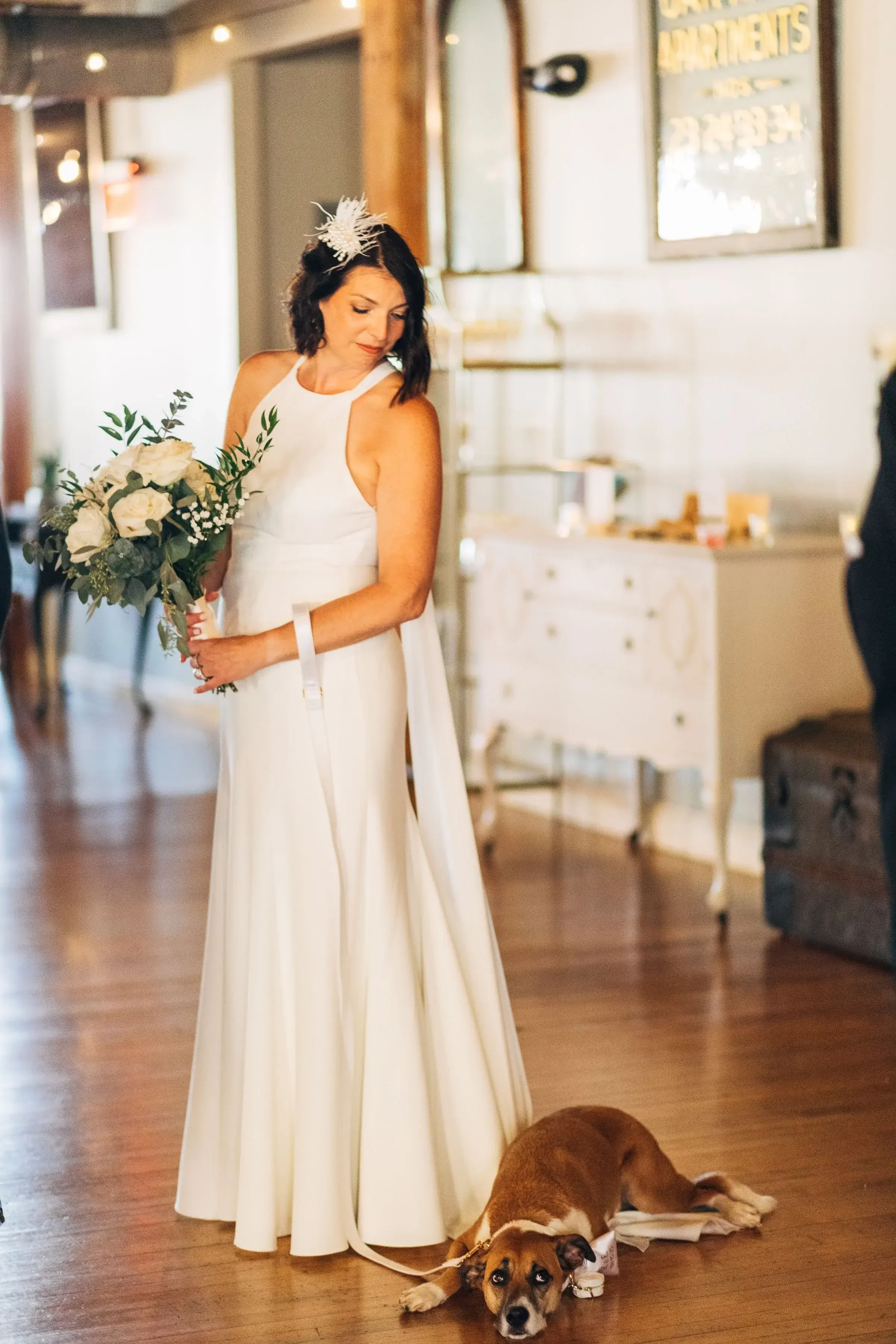 A woman in a white wedding dress holding a bouquet of white roses and greenery, standing indoors with a dog lying on the floor at her feet.