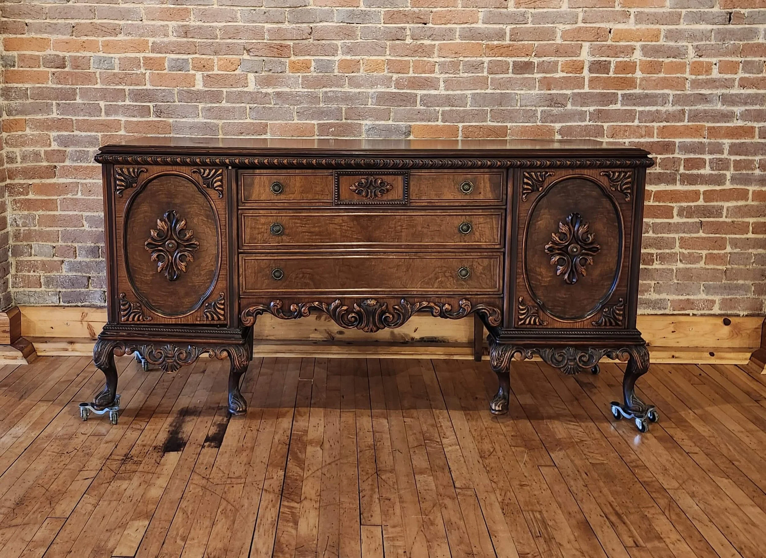 A vintage wooden sideboard with ornate carvings, set against an exposed brick wall and resting on a wooden floor.