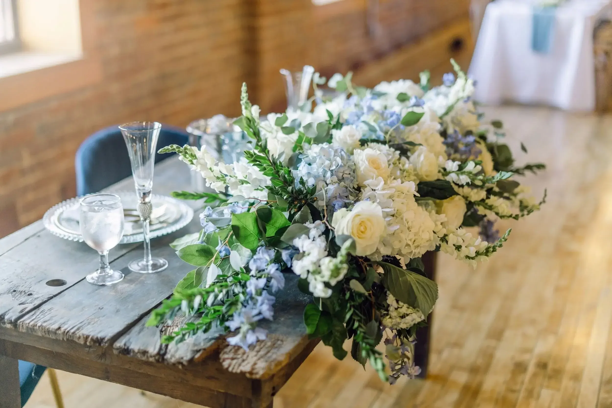 A rustic wooden table decorated with a large floral centerpiece of white and soft purple flowers along with green leaves, set for a celebration or wedding, with empty glassware and plates.