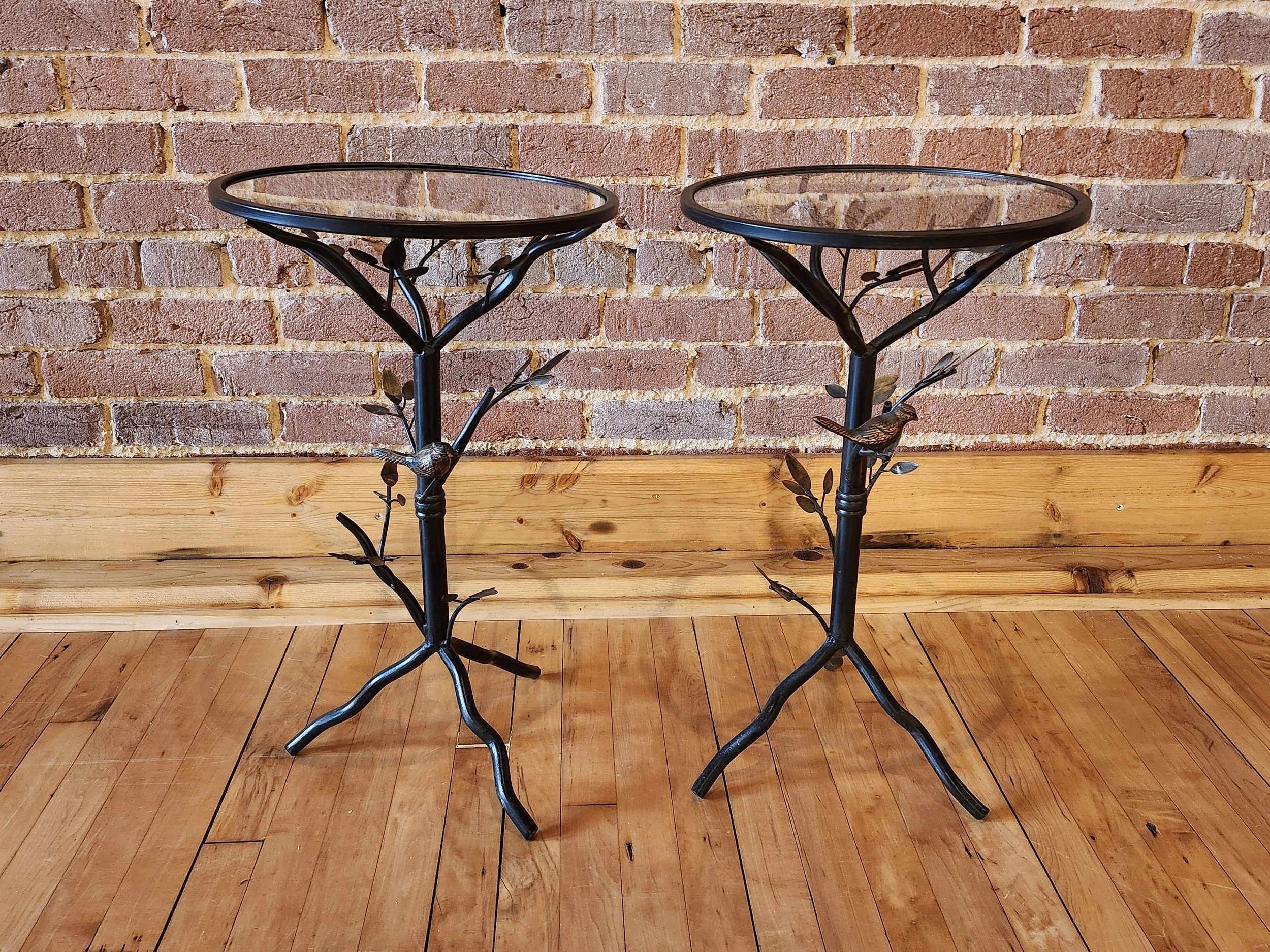 Two black metal plant stands with glass tops, decorative bird and branch designs, placed on a wooden floor against a brick wall background.
