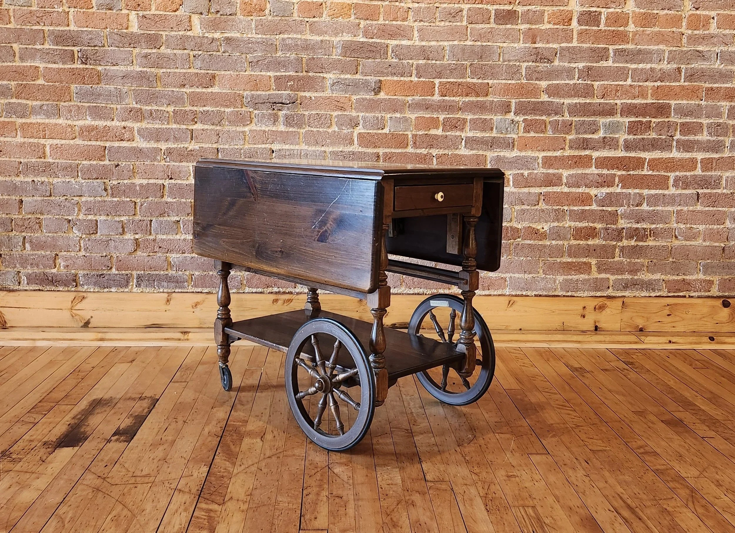 An antique wooden cart with wheels and a small drawer, placed against a brick wall with wooden flooring.