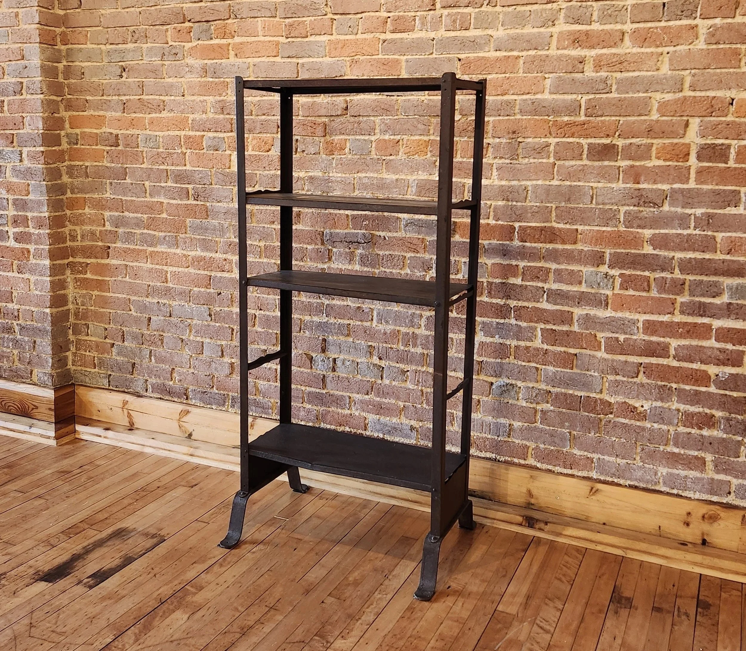 A black metal bookshelf with three wooden shelves, standing against a brick wall and on a wooden floor.