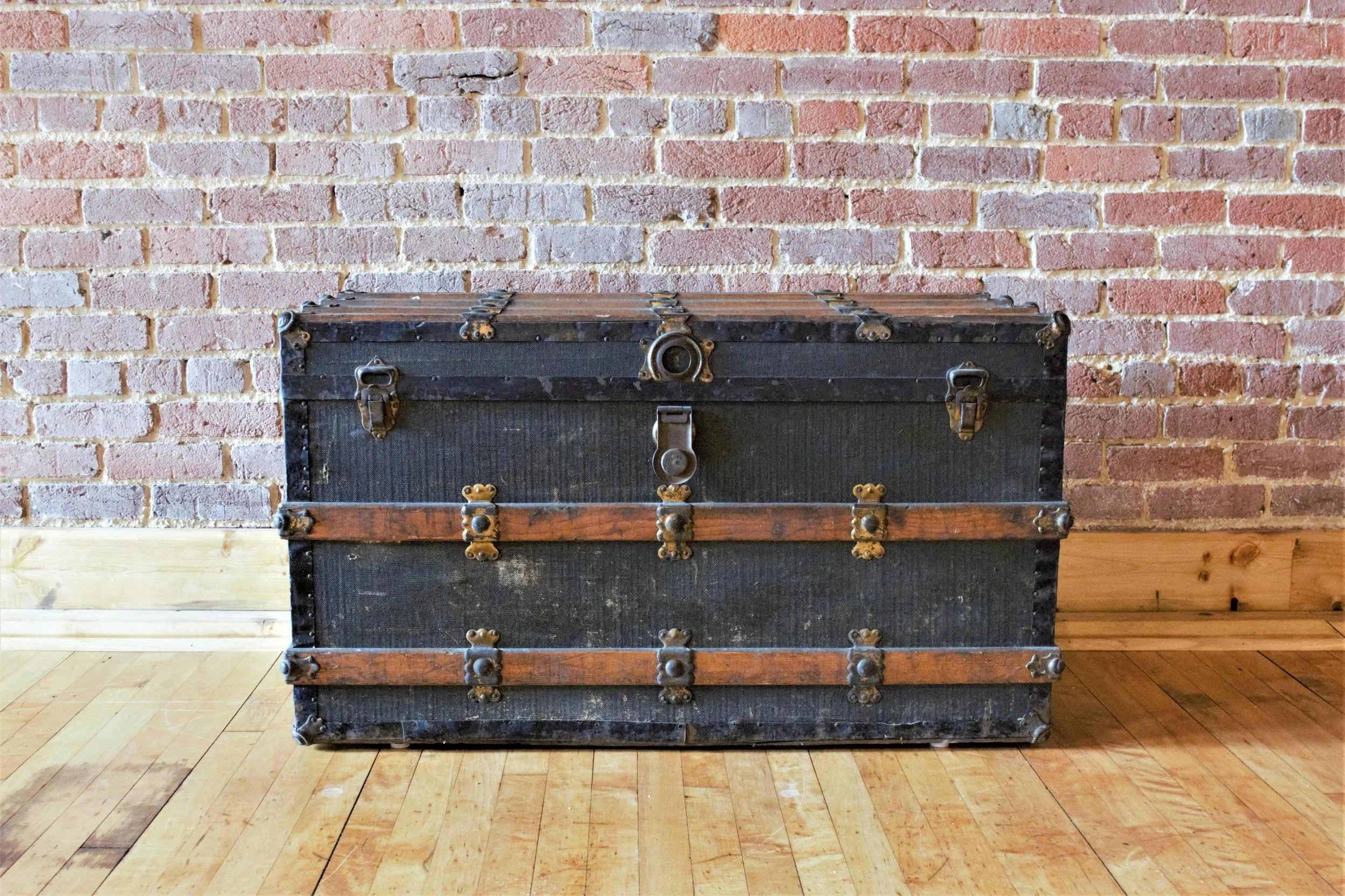 An antique black trunk with metal accents and wood details, placed on a hardwood floor against a brick wall.