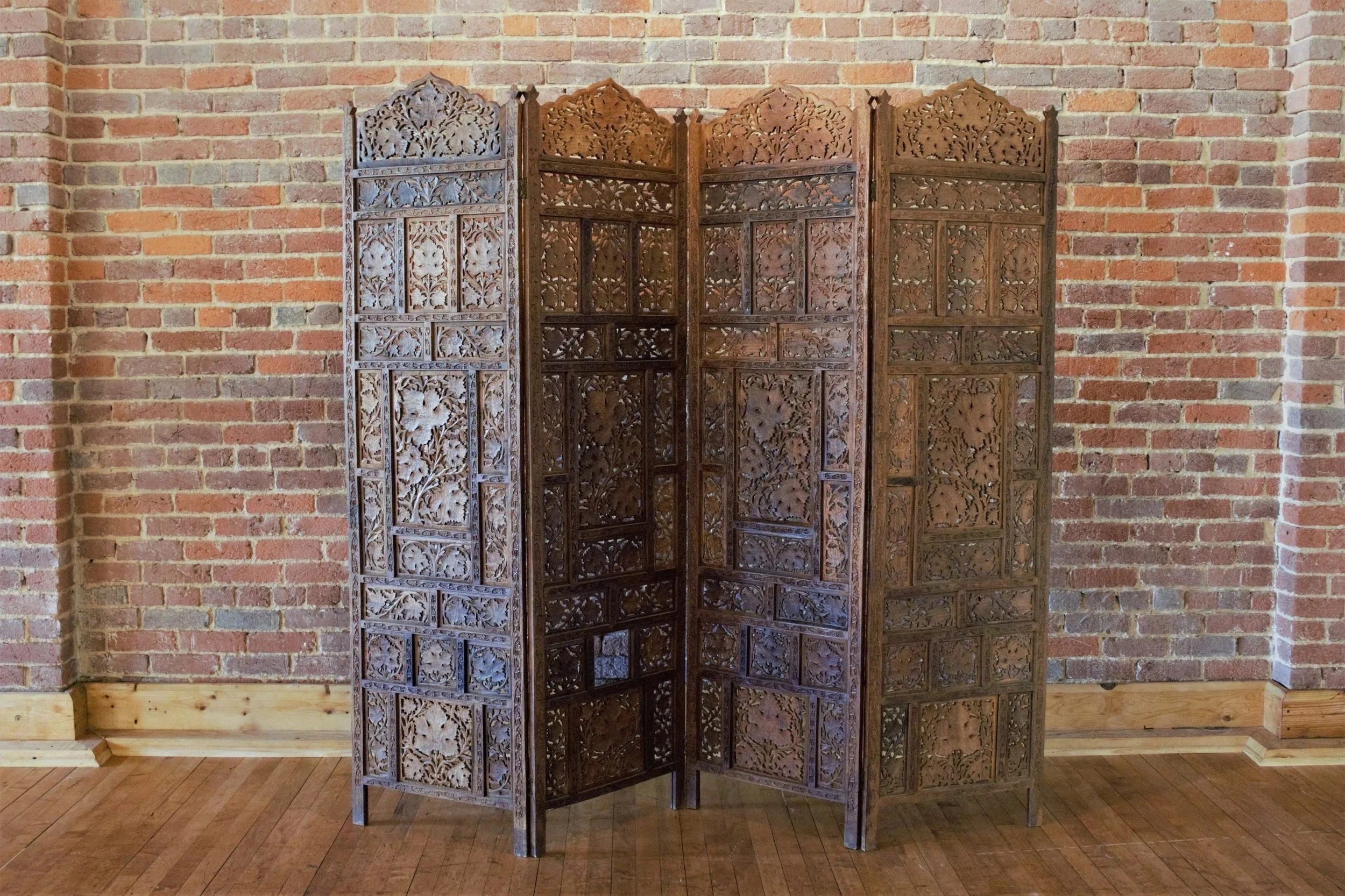 A wooden decorative folding room divider with intricate floral carvings, set against a brick wall and a wooden floor.