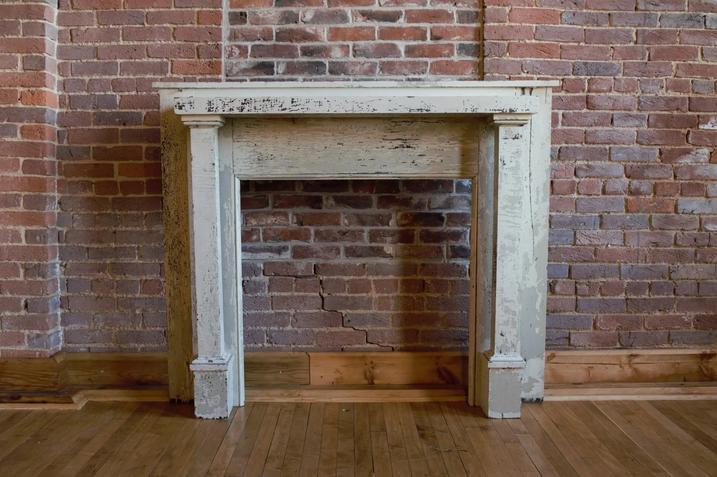 Empty vintage fireplace with chipped white paint, against a red brick wall in a room with wooden floor.