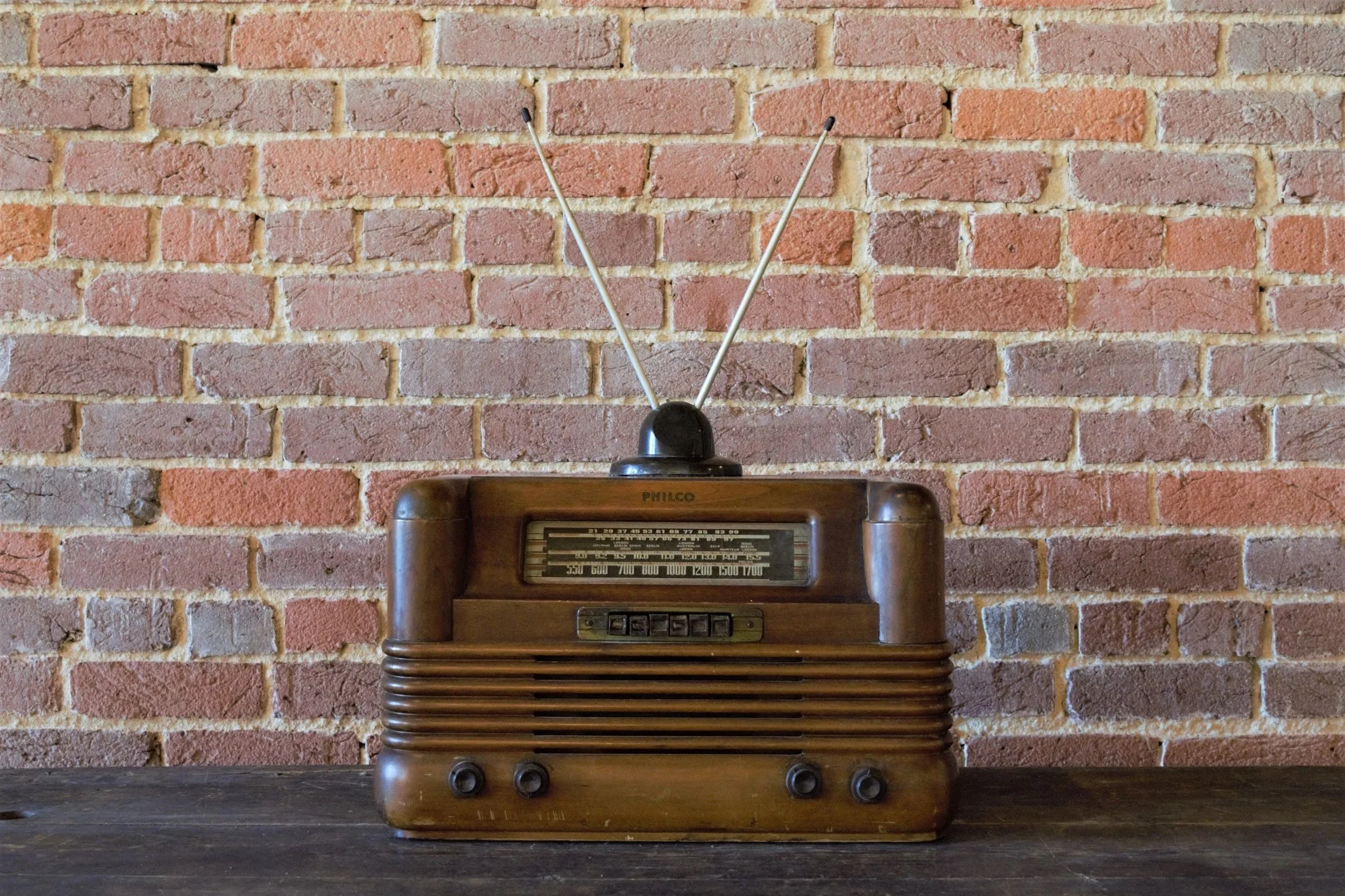A vintage wooden tabletop radio with antennas against a brick wall background.