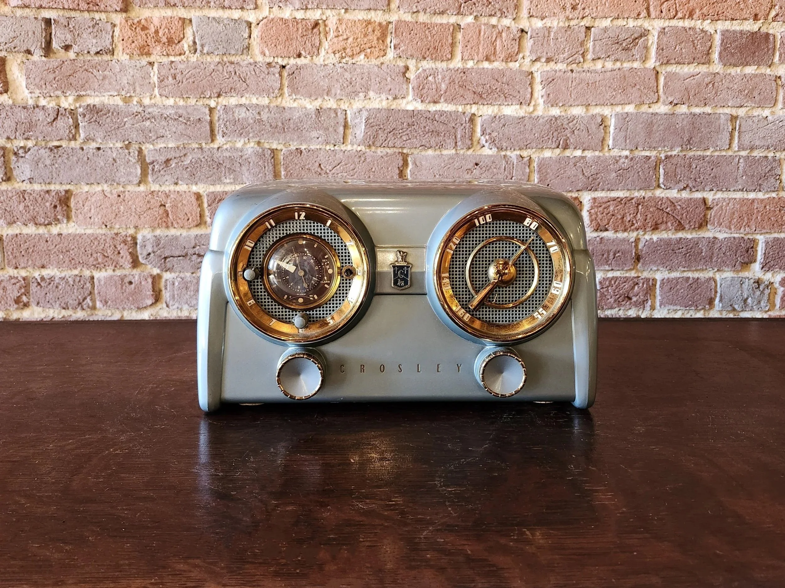A vintage Crosley radio with two dials, one for time and one for adjusting volume or station, placed on a dark wooden surface against a brick wall background.