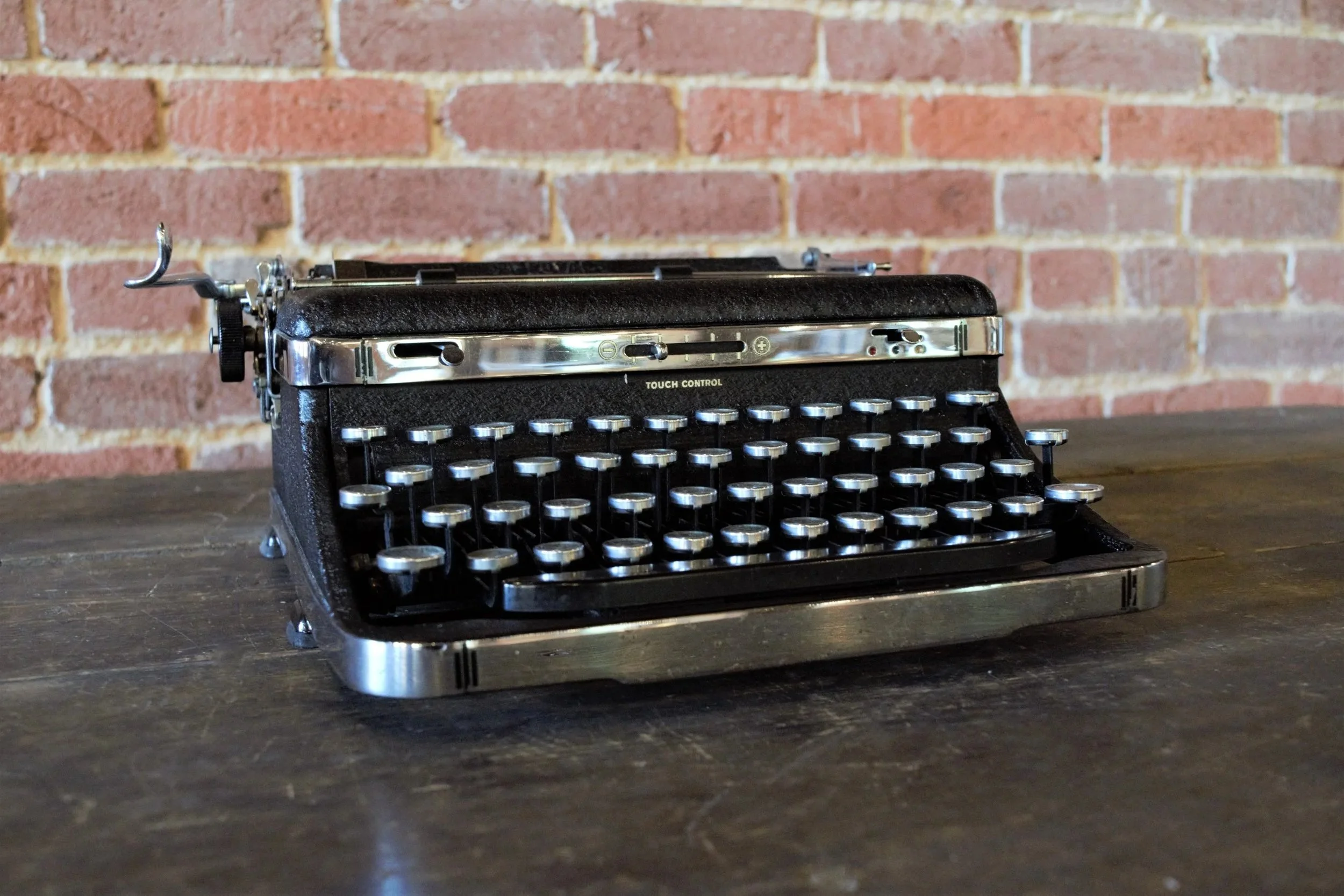 Vintage black typewriter with silver accents on a wooden surface against a red brick wall.