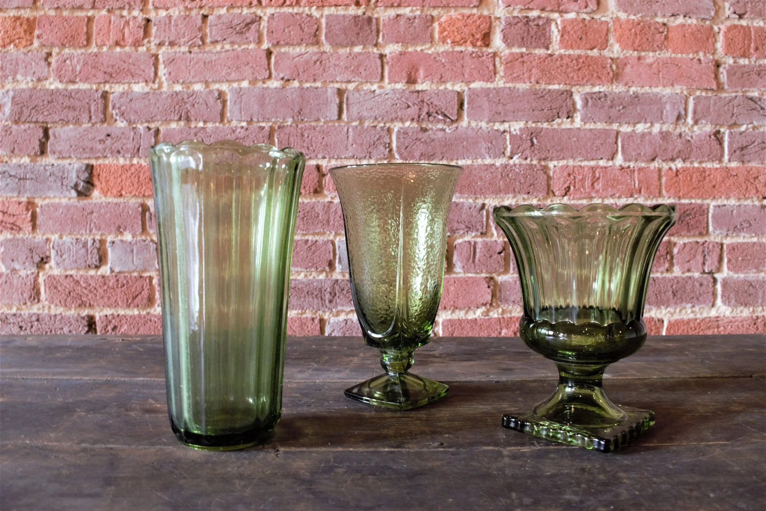 Three green glass vases of different shapes and sizes on a wooden table against a pink brick wall.