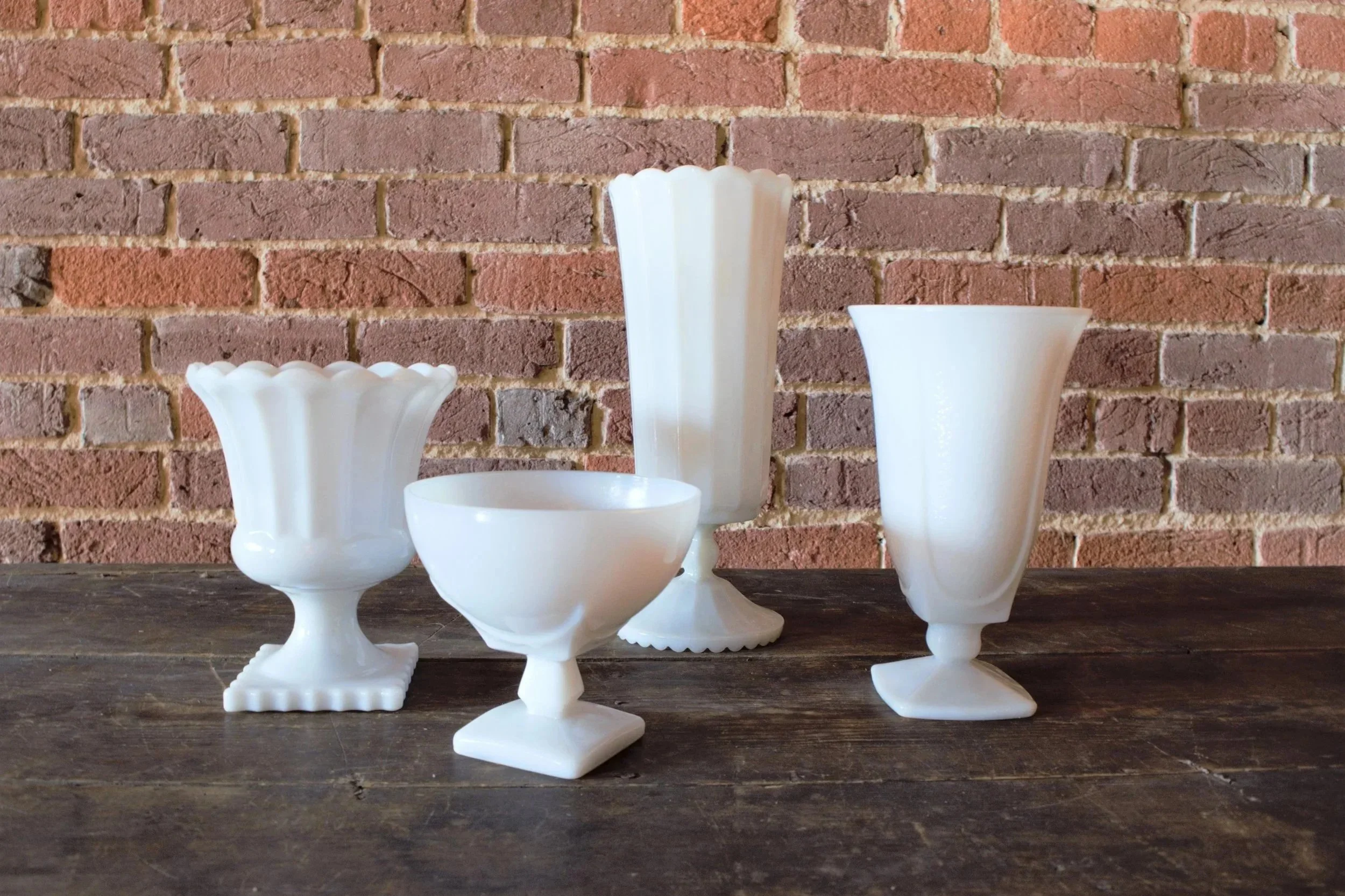 Five white milk glass vases with different shapes and sizes arranged on a rustic wooden table against a brick wall background.