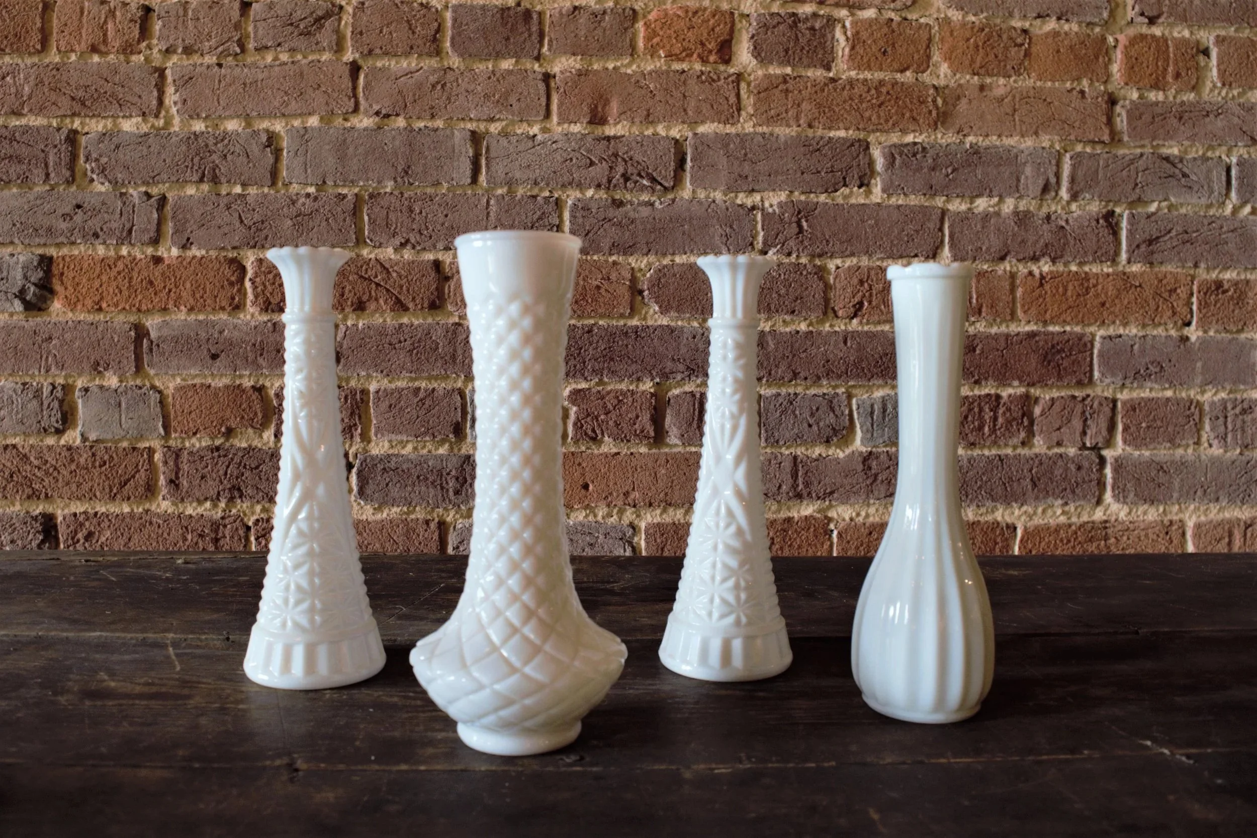 Four white decorative glass vases of various heights and patterns on a dark wooden surface in front of a red brick wall.