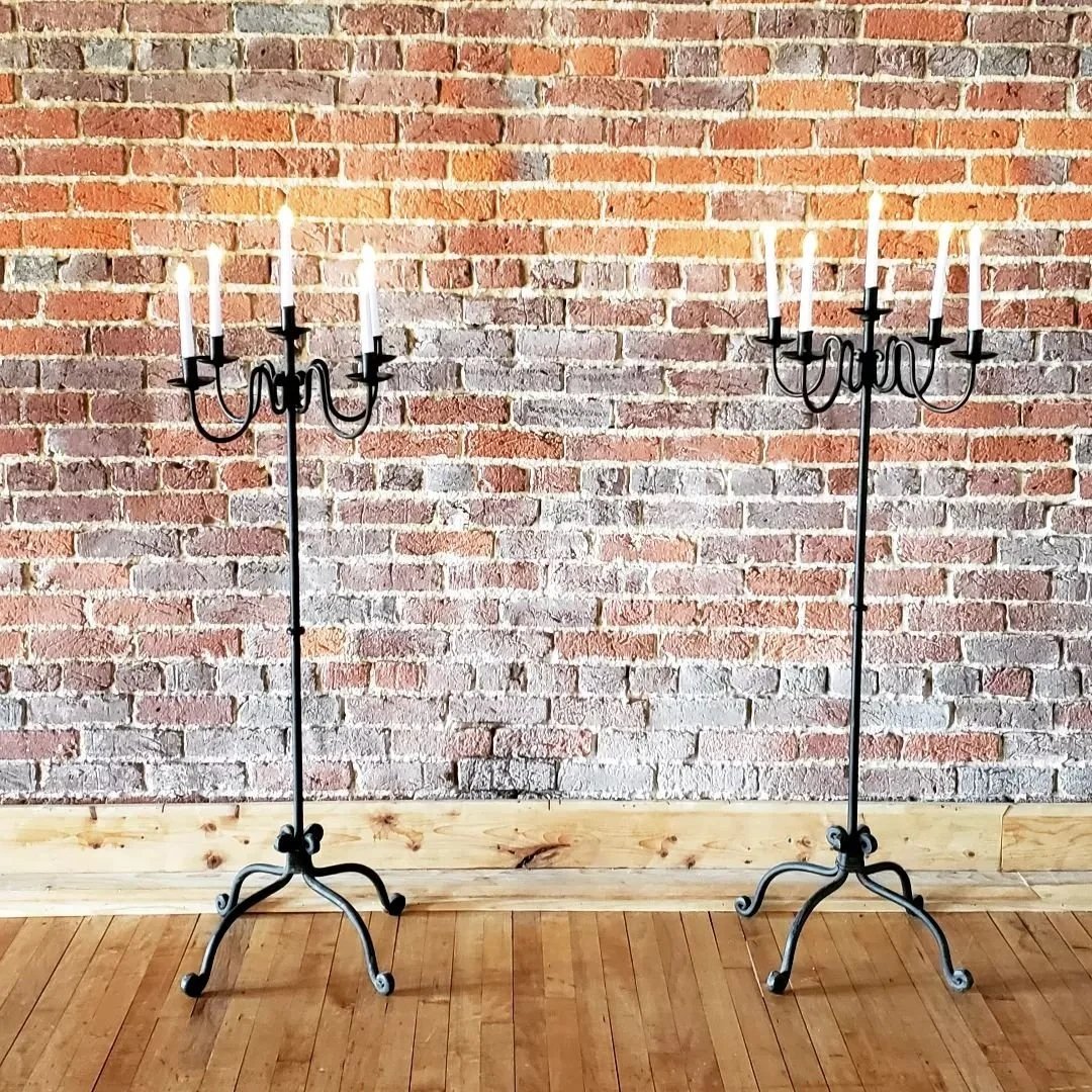 Two black metal candelabras with five white candles each, standing on a wooden floor against a brick wall.