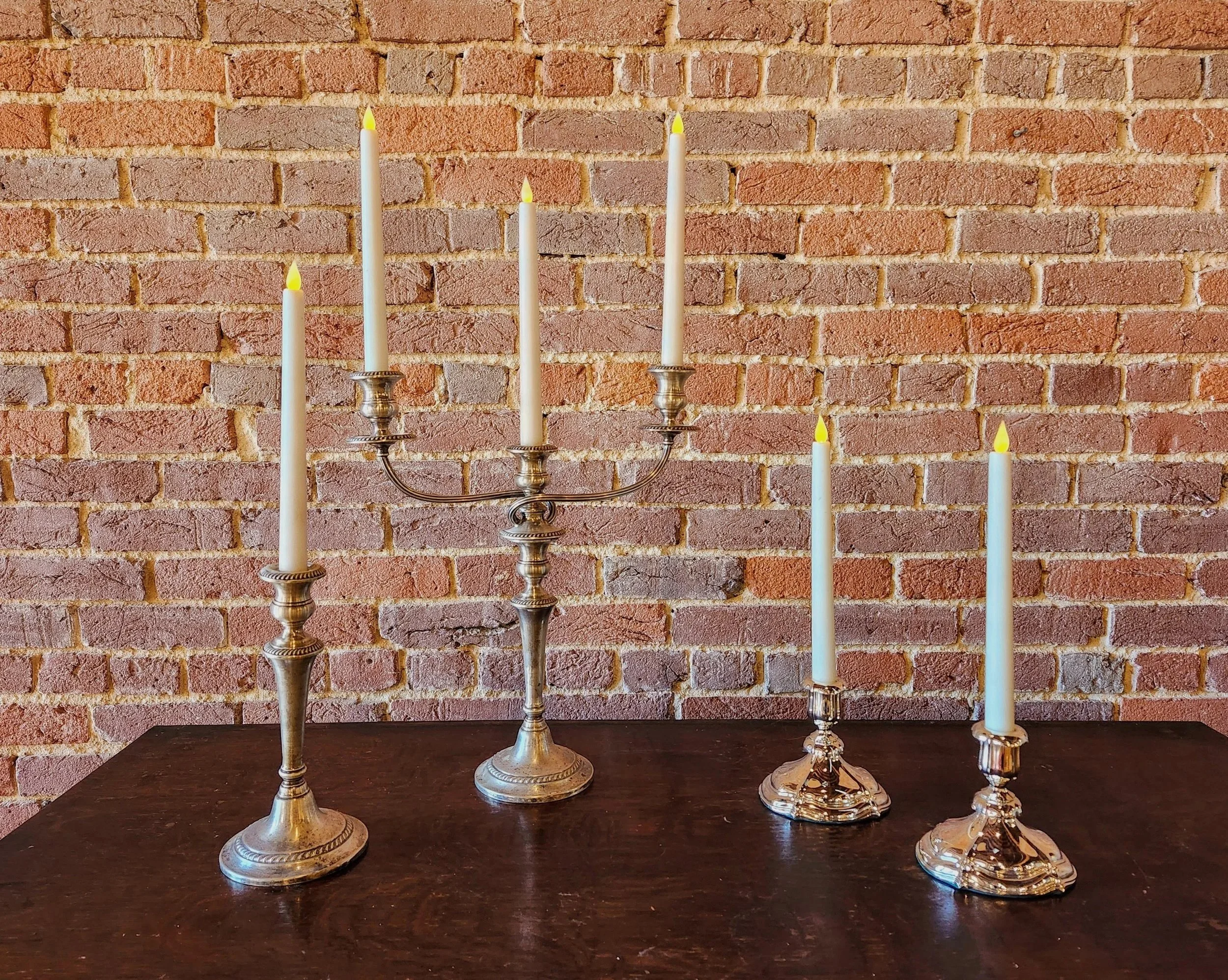 Four lit candles in silver candleholders are placed on a dark wooden table against a red brick wall.