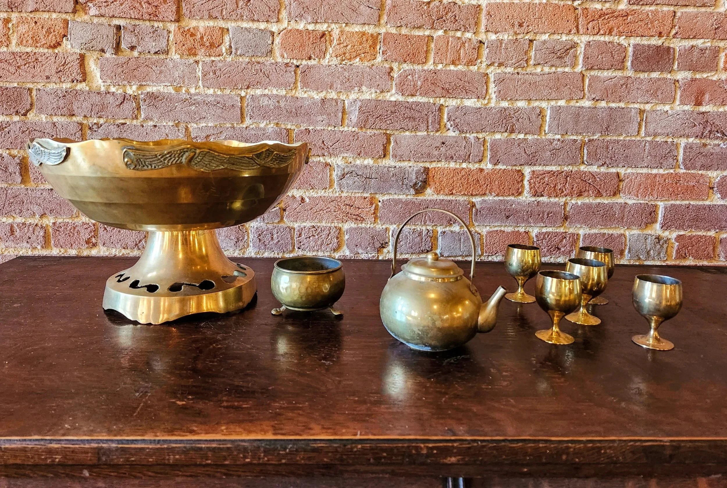 A brass tea set with a teapot and six small cups on a wooden table, with a large brass bowl on the left and a small brass bowl in the middle, against a brick wall background.