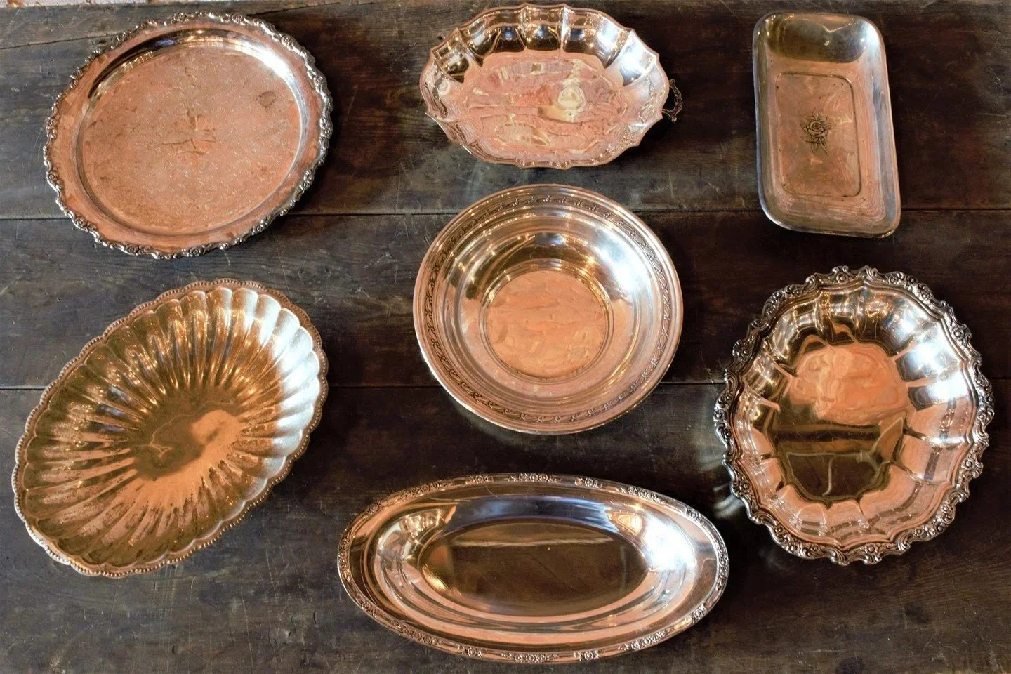 Set of seven antique silver or silver-plated trays and bowls with ornate edges, arranged on a dark wooden table.