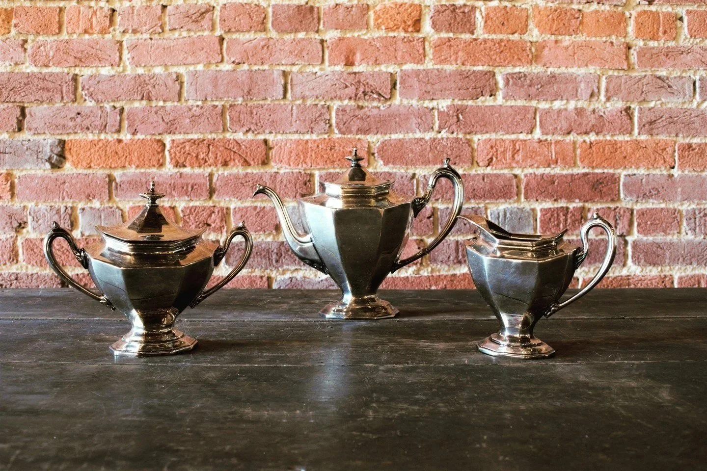 Three silver tea sets with teapots and sugar bowls, placed on a dark wooden surface in front of a red brick wall.