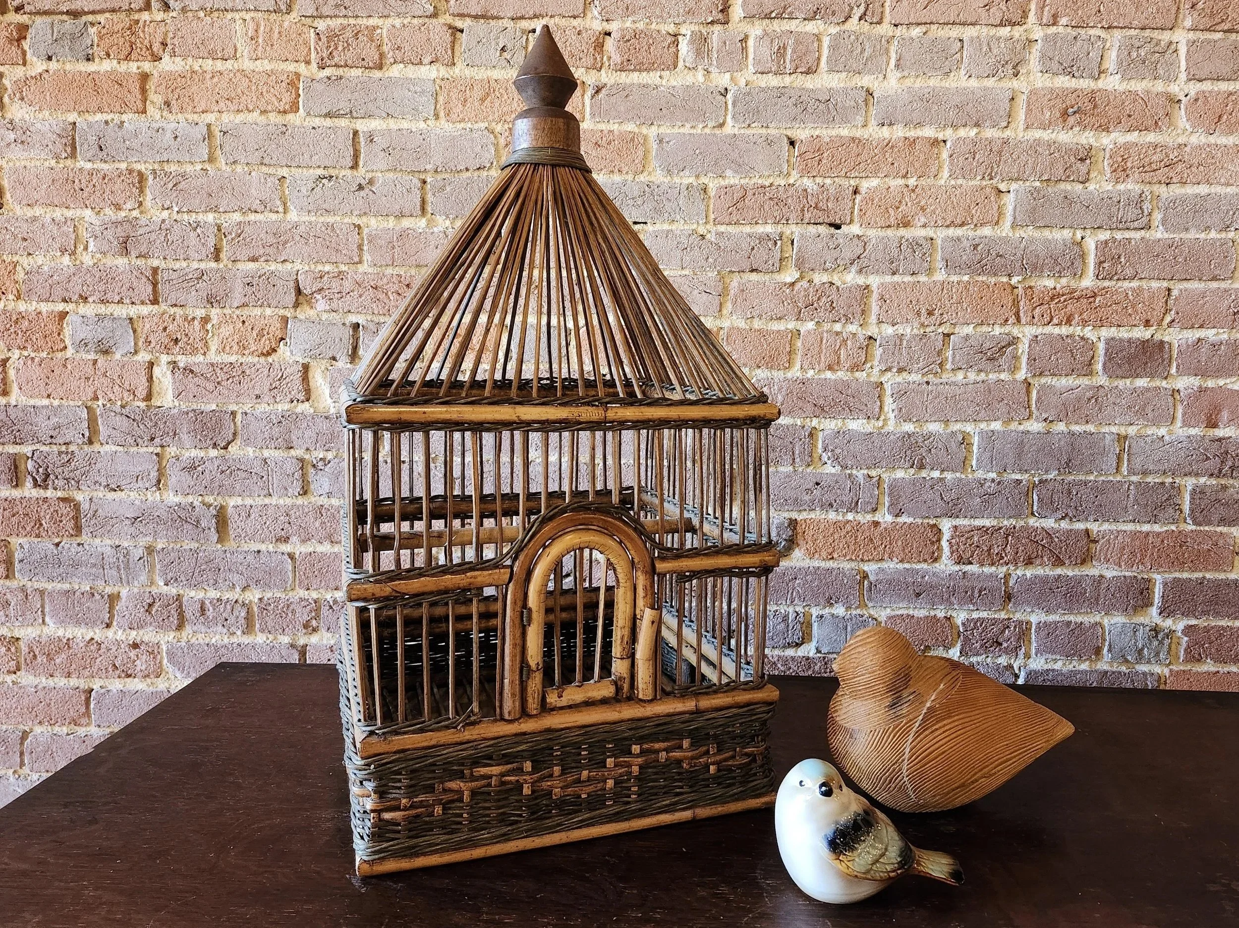 Decorative birdcage made of natural wicker and wood, placed on a dark wooden surface against a brick wall, accompanied by a ceramic bird figurine and a large wooden leaf-shaped object.