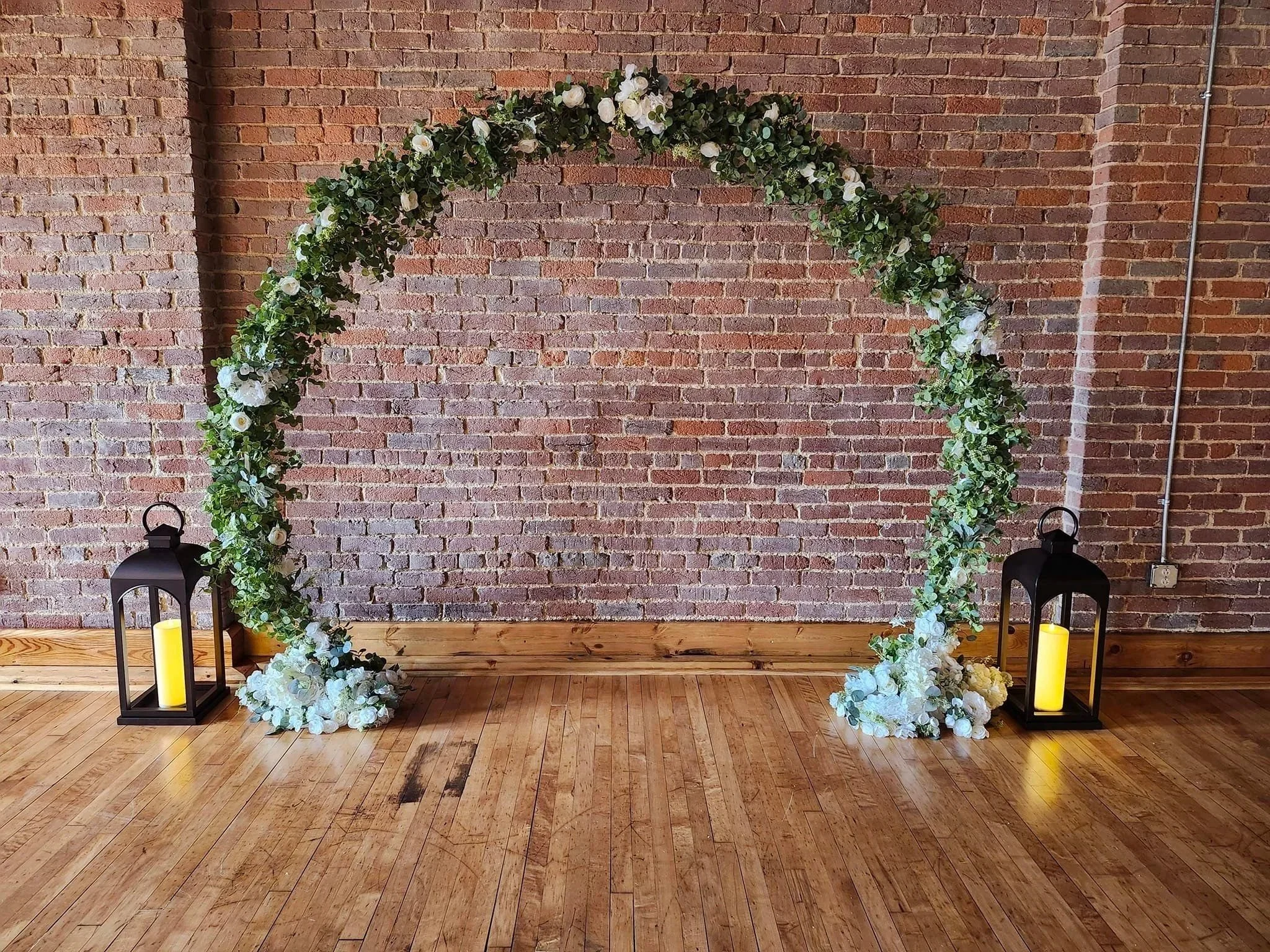 A floral wedding arch decorated with white and green flowers, two black lanterns with yellow candles on the floor flanking the arch, against a red brick wall and wooden floor.