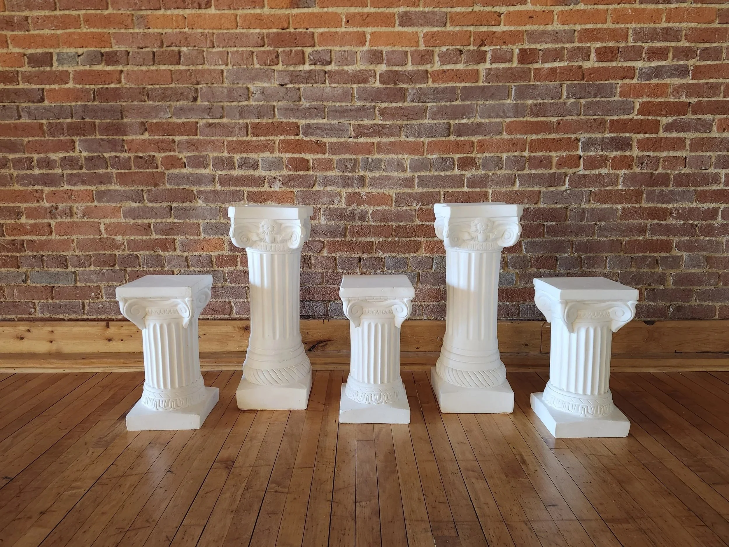 Five white classical style pedestal columns arranged against a brick wall with a wooden floor.