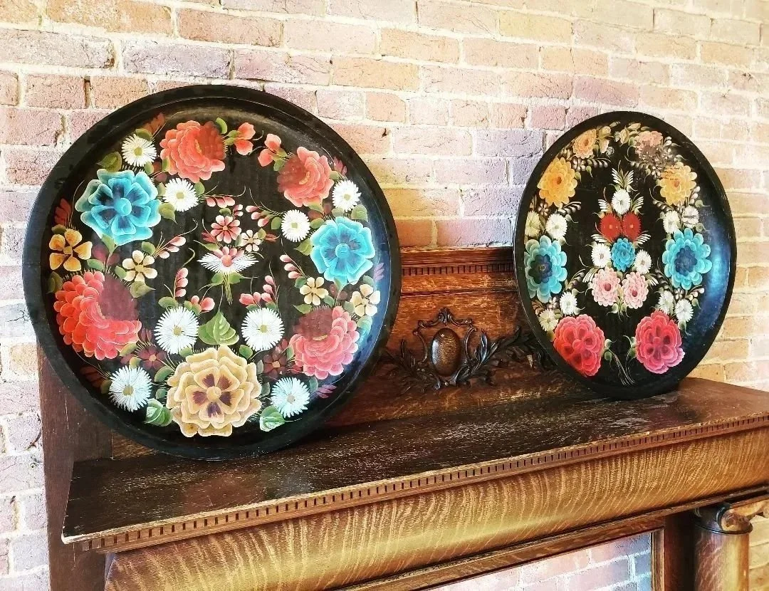Two decorative floral trays with vibrant painted flowers, placed on a wooden shelf against a brick wall.