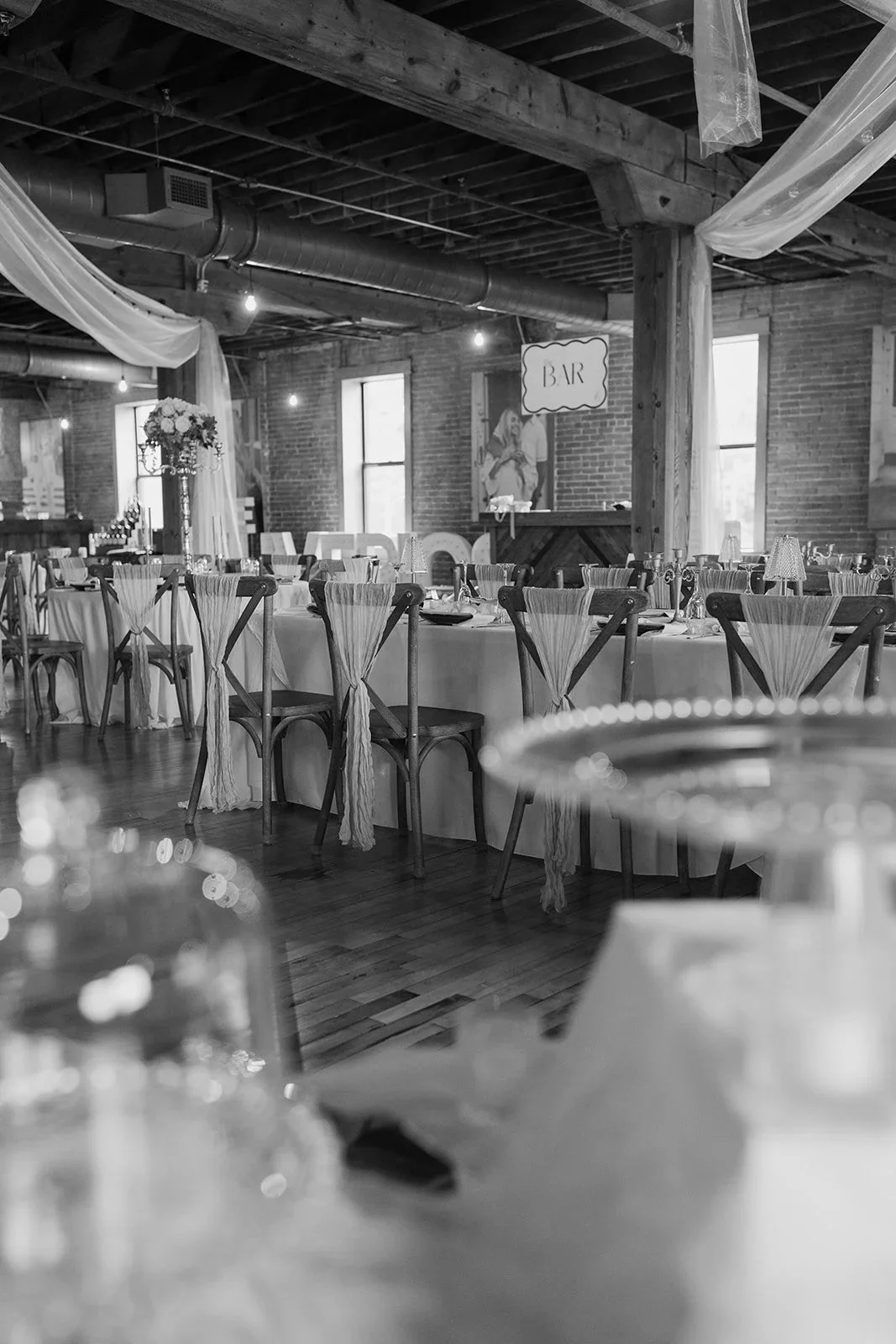 Black and white photo of a decorated event space with long tables, chairs with streamers, a bar sign, and floral arrangements, in a rustic venue with exposed brick walls and wooden beams.