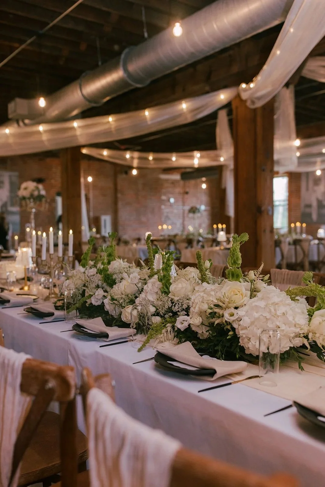 Long banquet table with white tablecloth decorated with a large floral arrangement of white flowers and greenery, set for a formal event in a rustic venue with exposed brick walls, wooden beams, and string lights.