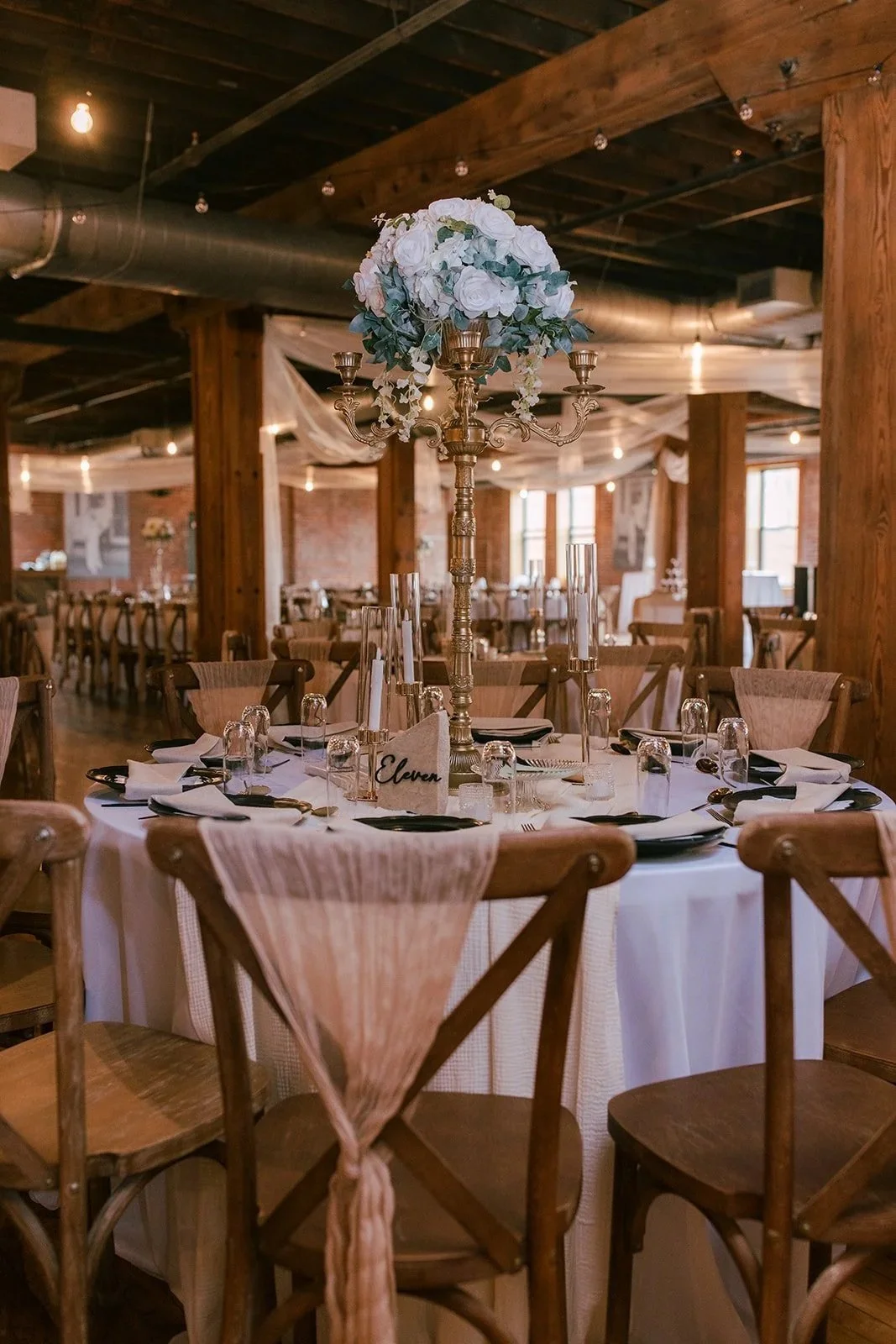 Elegant wedding or event reception table with a white tablecloth, black plates, gold utensils, tall glass candle holders, and a tall floral centerpiece with white flowers and greenery, set in a rustic wooden hall.