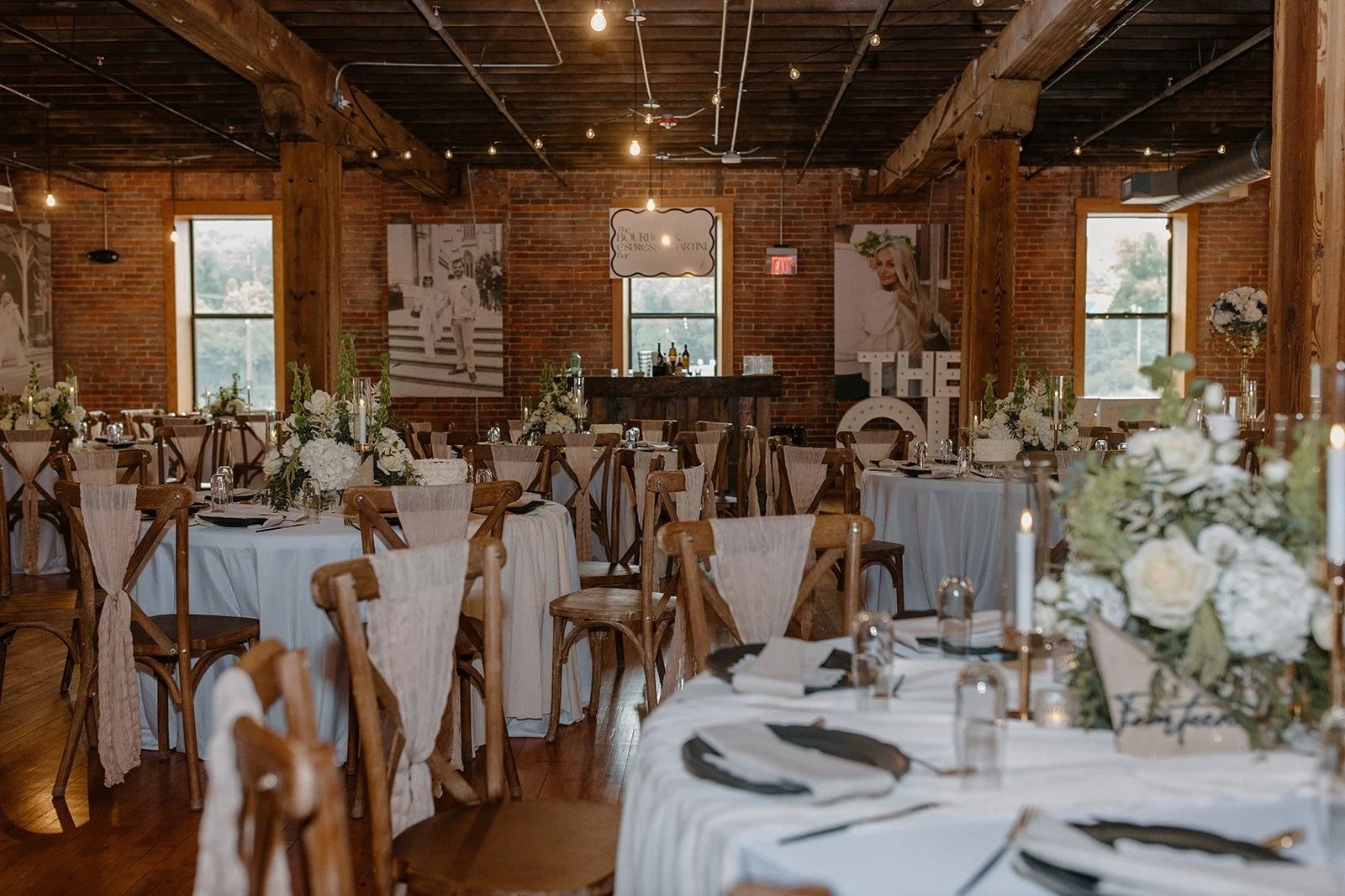 Indoor wedding reception decorated with white flowers, candles, and black napkins, set on round tables with wooden chairs, in a rustic brick and wood interior.