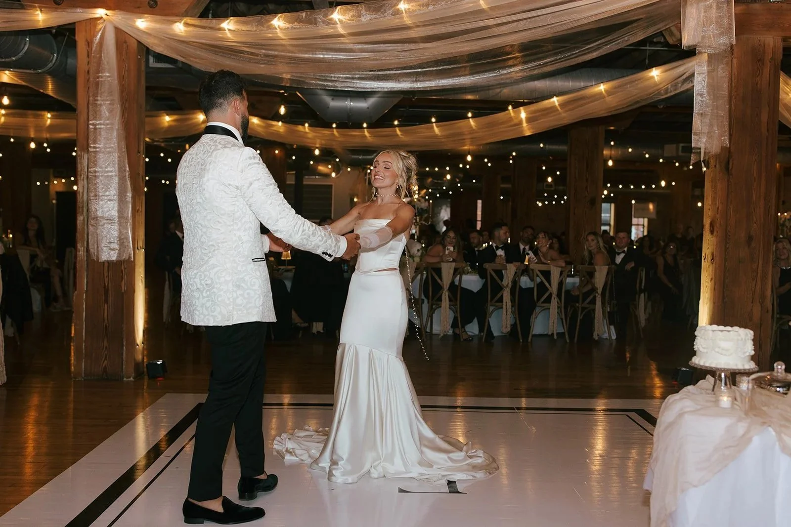 Bride and groom dancing at their wedding reception with guests seated at tables in the background, decorated with fairy lights and sheer fabric draping in a rustic venue.