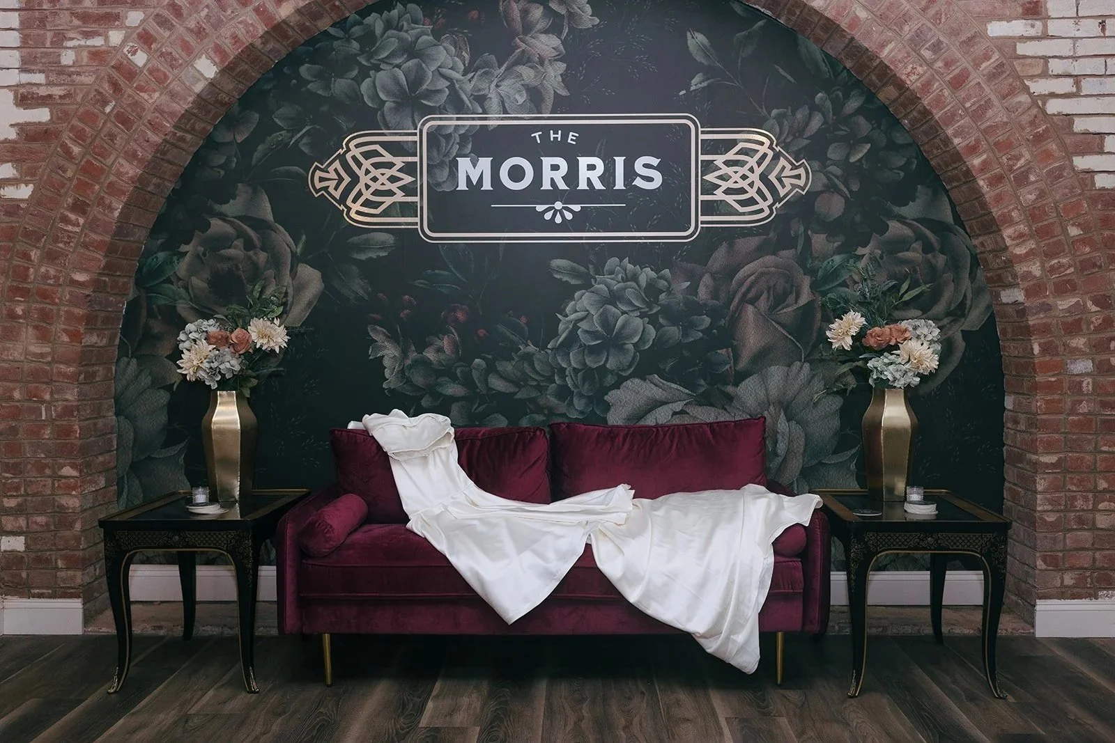 Velvet maroon sofa with white draped fabric and two floral arrangements on black side tables against a dark floral mural with the logo "The Morris" on a brick wall background.
