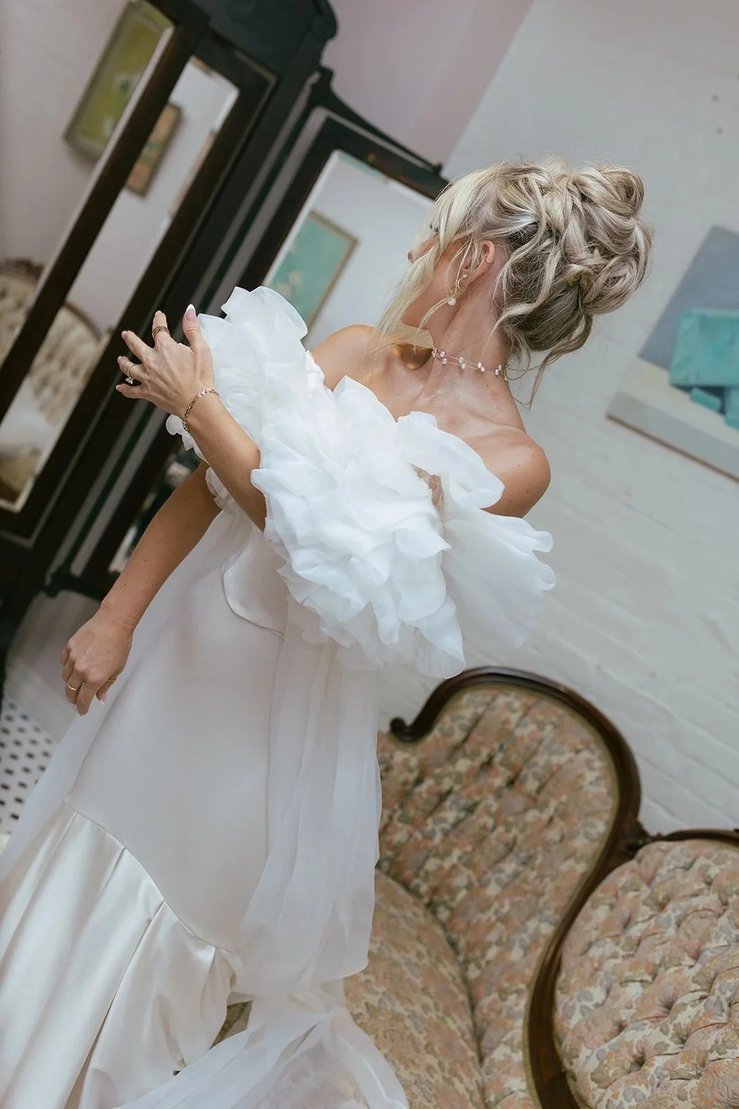 A woman in a white dress with ruffled sleeves and a pearl necklace, with hair styled in an elaborate updo, standing in a room with vintage furniture, looking away from the camera.