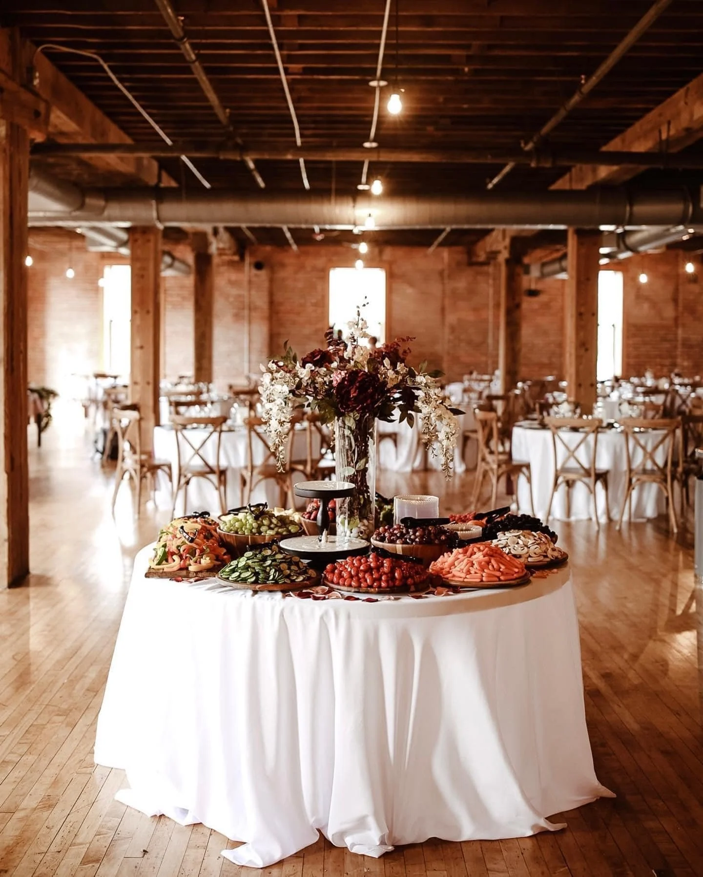 A table with a flower centerpiece and various foods, including grapes, strawberries, cookies, and sliced vegetables, set in a rustic banquet hall with wooden beams and round tables.