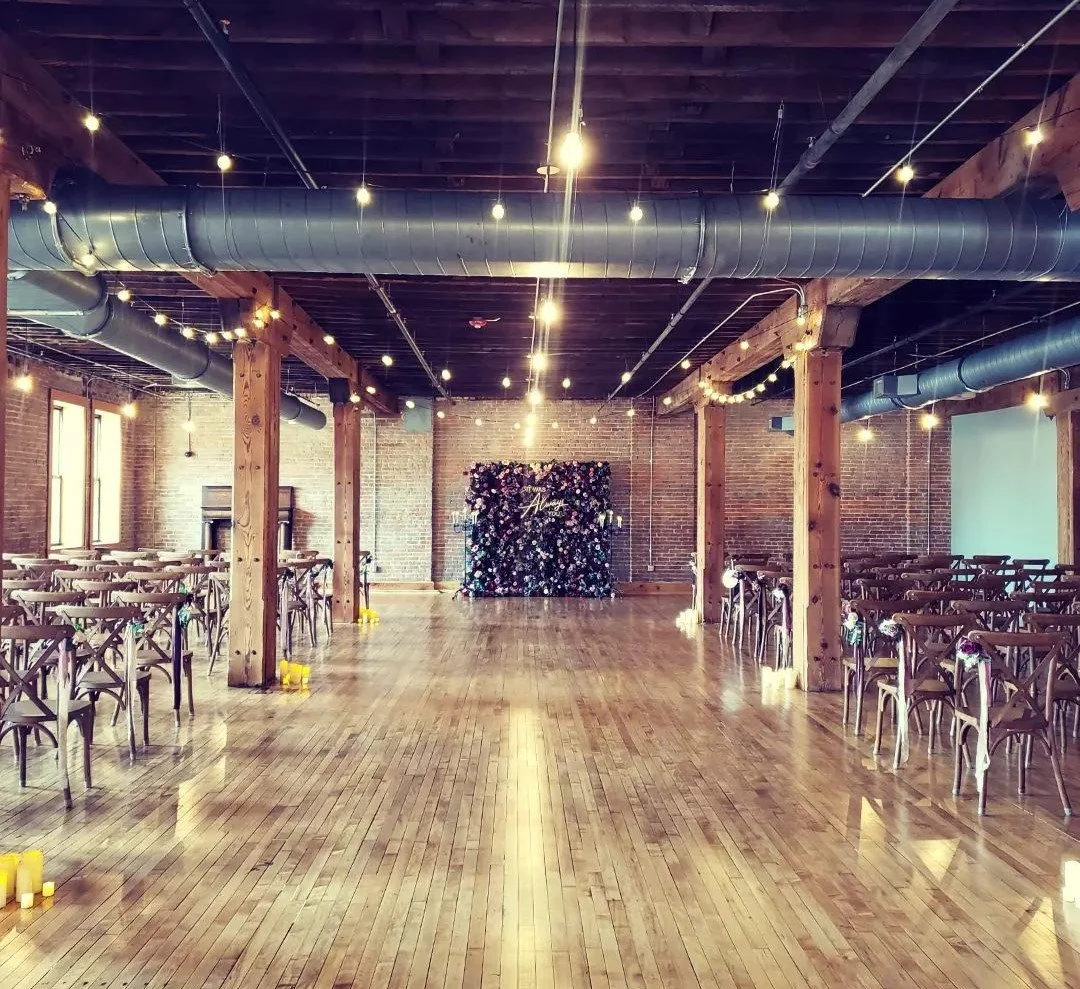An event space with a polished wooden floor, exposed brick walls, wooden beams, ceiling ductwork, and string lights hanging overhead. There are tables and chairs set up on either side of the room, and a floral backdrop with the word 'Always' in the d