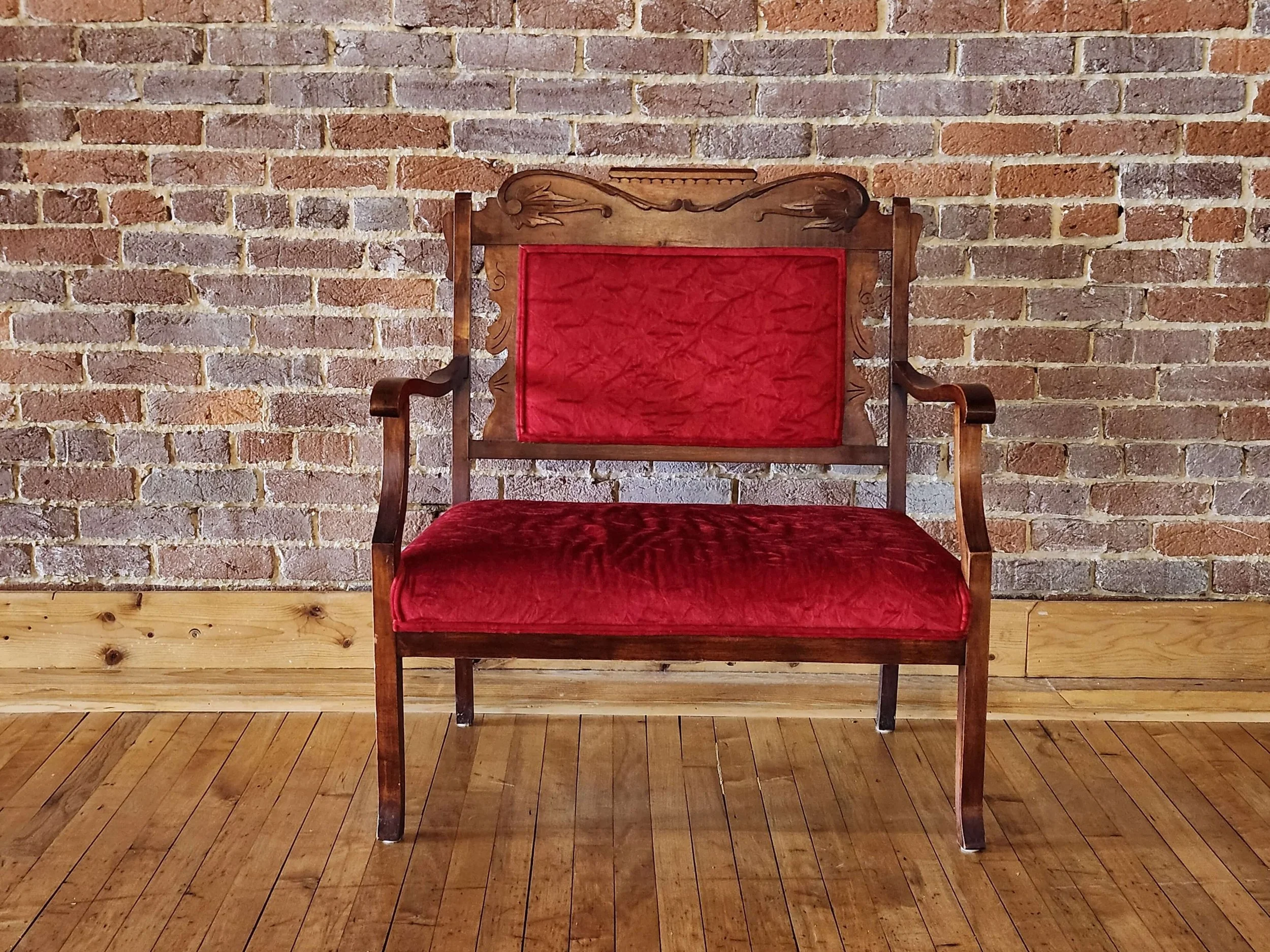 An antique wooden bench with a red velvet cushion and backrest, positioned against a brick wall and wooden floor.