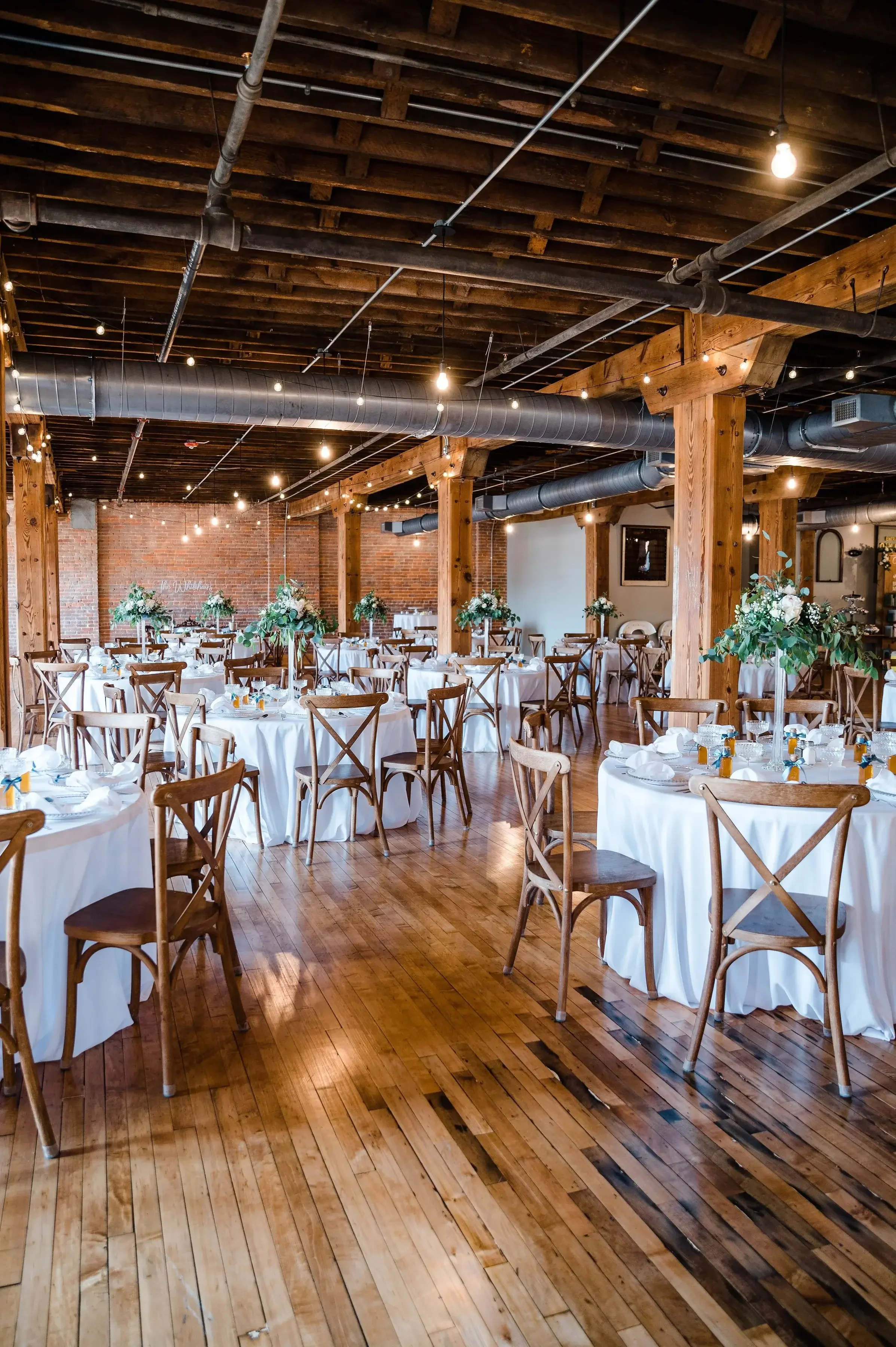 A spacious event hall decorated for a wedding reception with round tables covered in white tablecloths, adorned with floral centerpieces, set with plates and glasses, surrounded by wooden chairs, and illuminated with string lights hanging from the wo